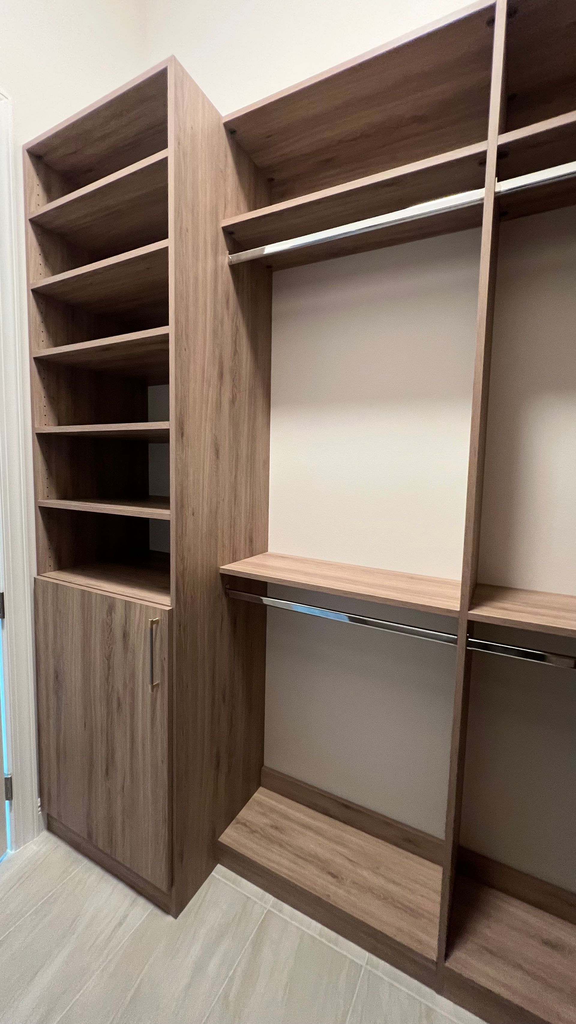 A light wood-grain walk-in closet system with open shelves, hanging rods, and a cabinet door.
