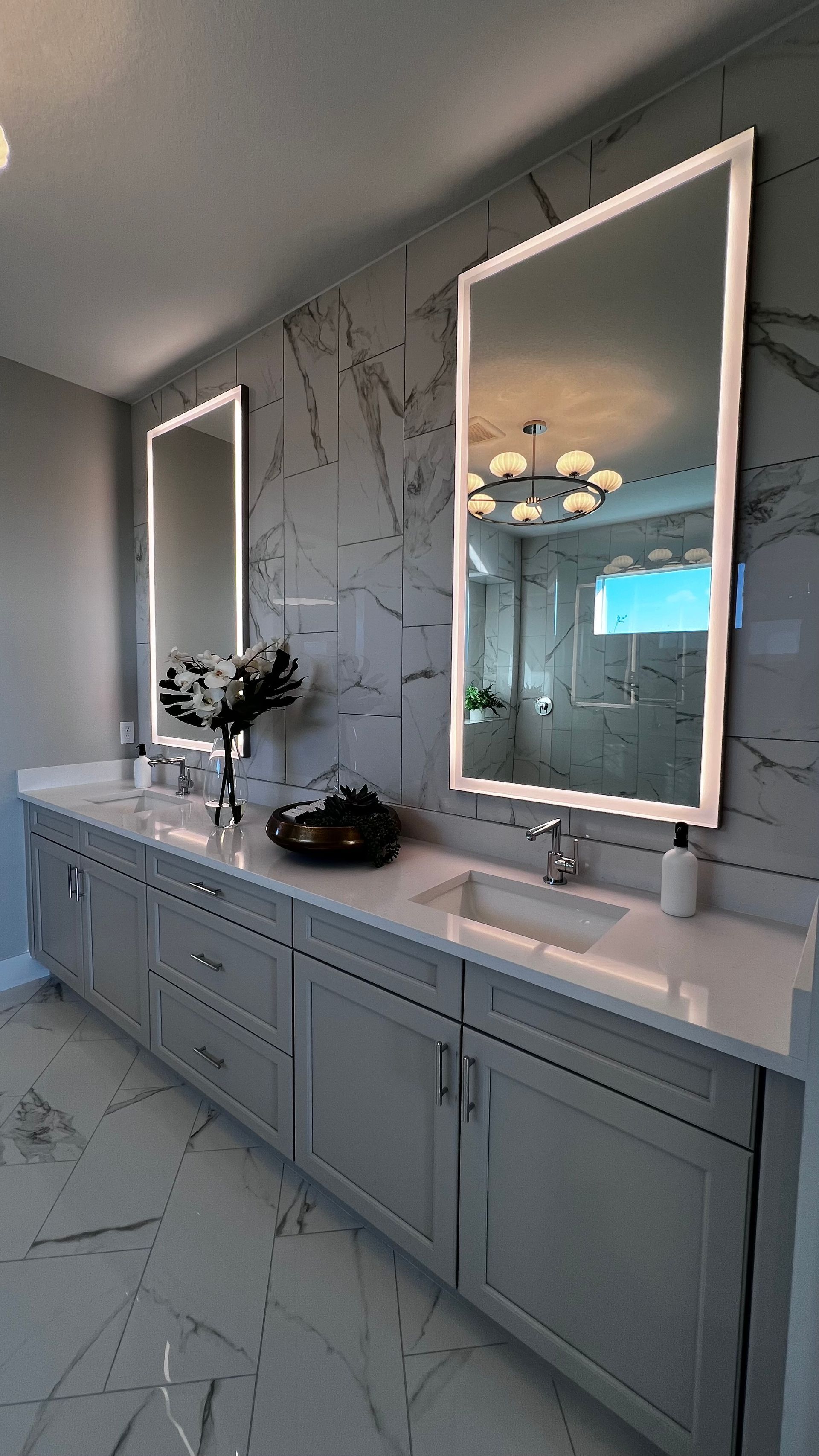 A modern bathroom with grey cabinets, white marble countertops, a wall with marble accents, and two backlit mirrors.
