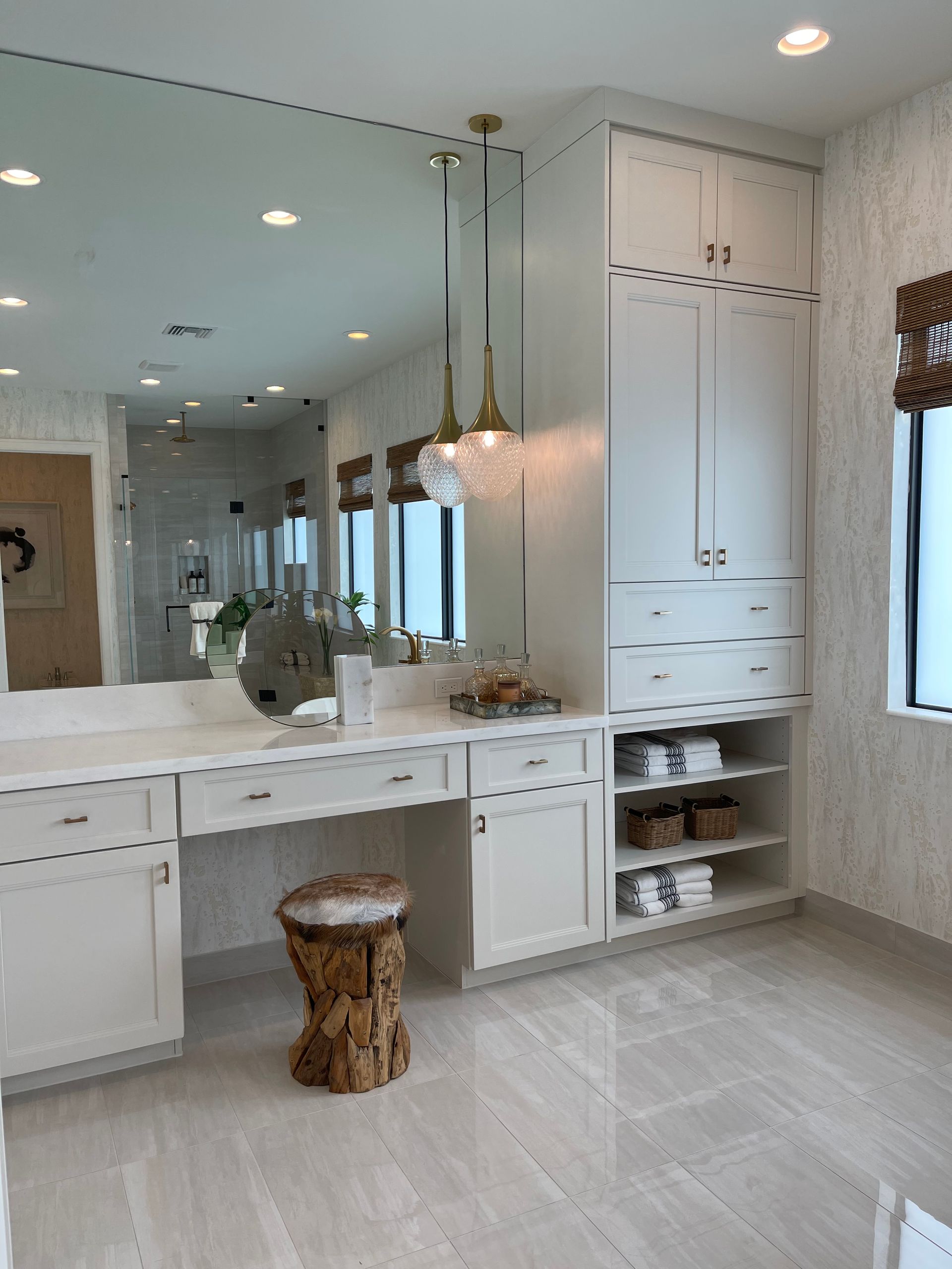 A white vanity with a mirror, storage cabinets, open shelving for towels, and a rustic wood stool in a bright bathroom.