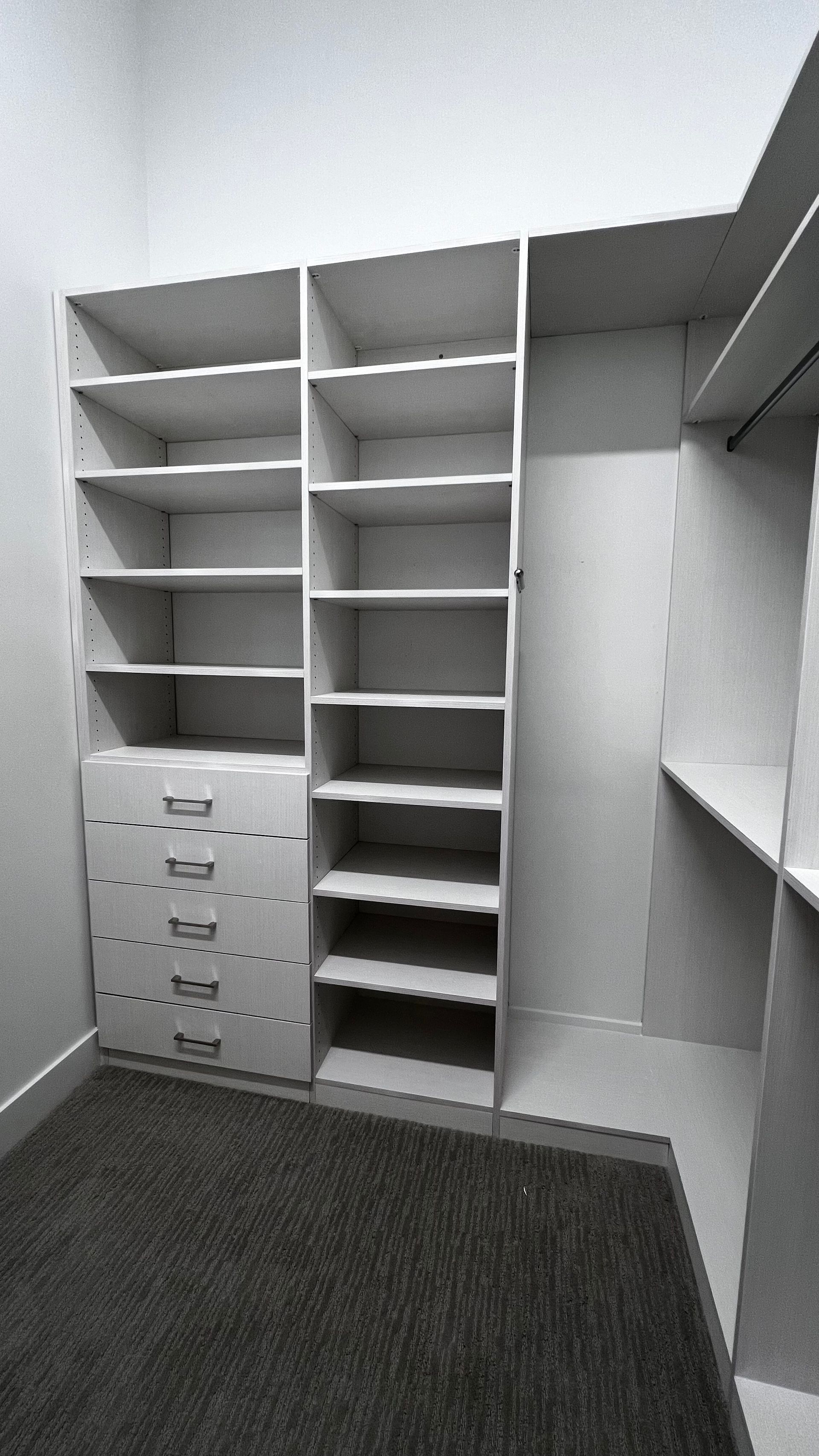 A walk-in closet featuring white wooden shelving units with an integrated five-drawer dresser on a dark carpet floor.