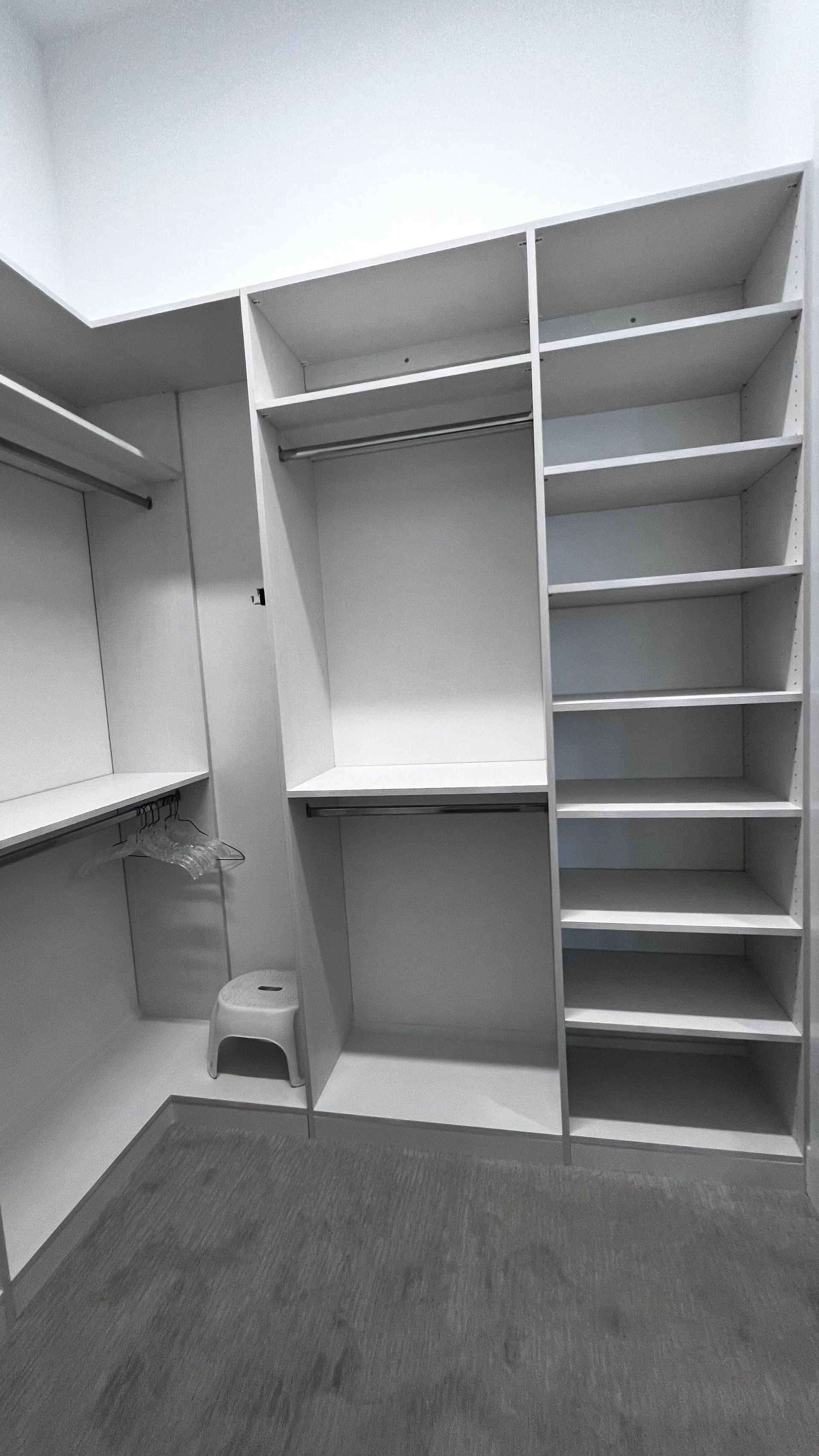 An empty white walk-in closet with shelves, hanging rods, and a small stool on a gray carpeted floor.