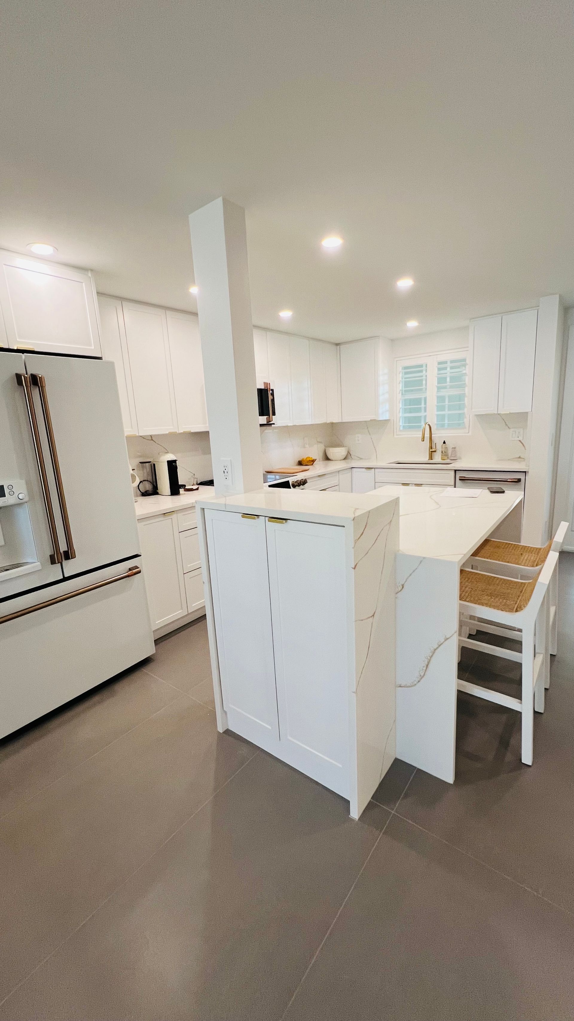 A modern, brightly lit kitchen featuring white cabinets, a central island with bar stools, and a white refrigerator.