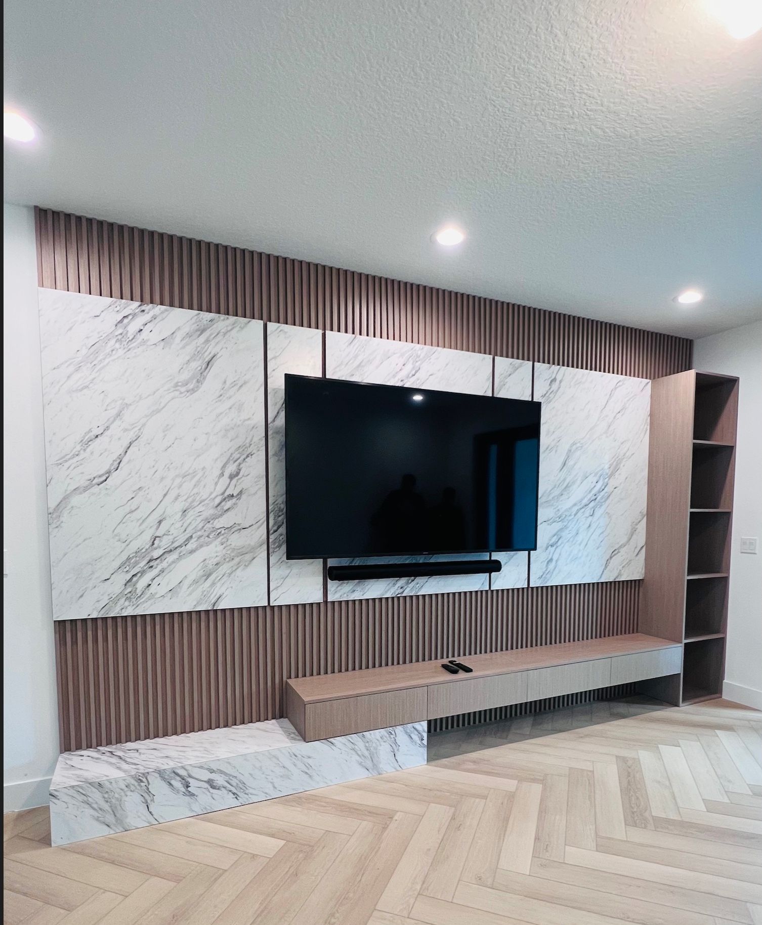 A wall-mounted TV framed by marble panels and wood slat detailing, featuring a floating cabinet and a tall side shelf.