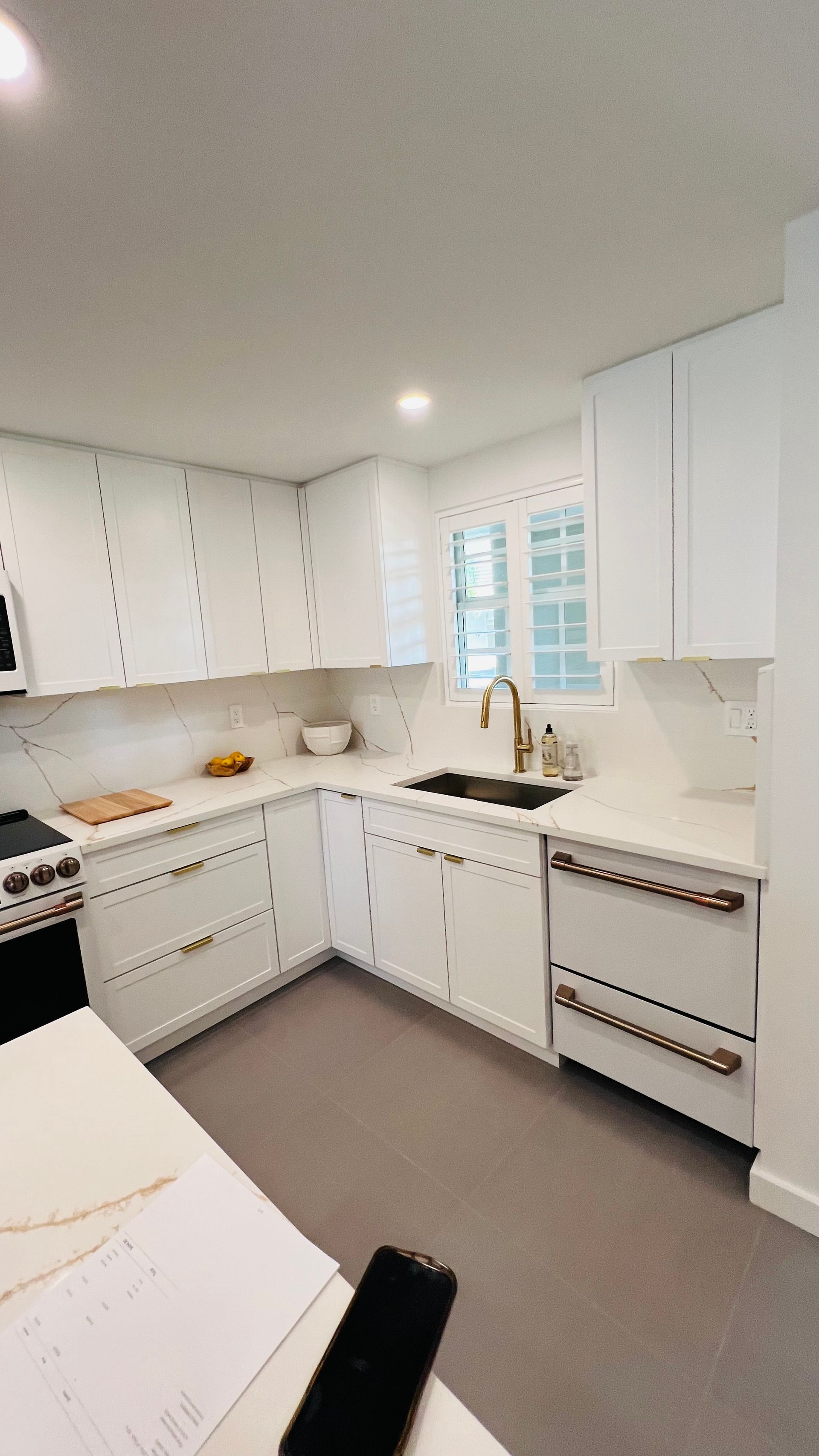 A white L-shaped kitchen with marble countertops, gold hardware, a black sink, and light gray flooring.