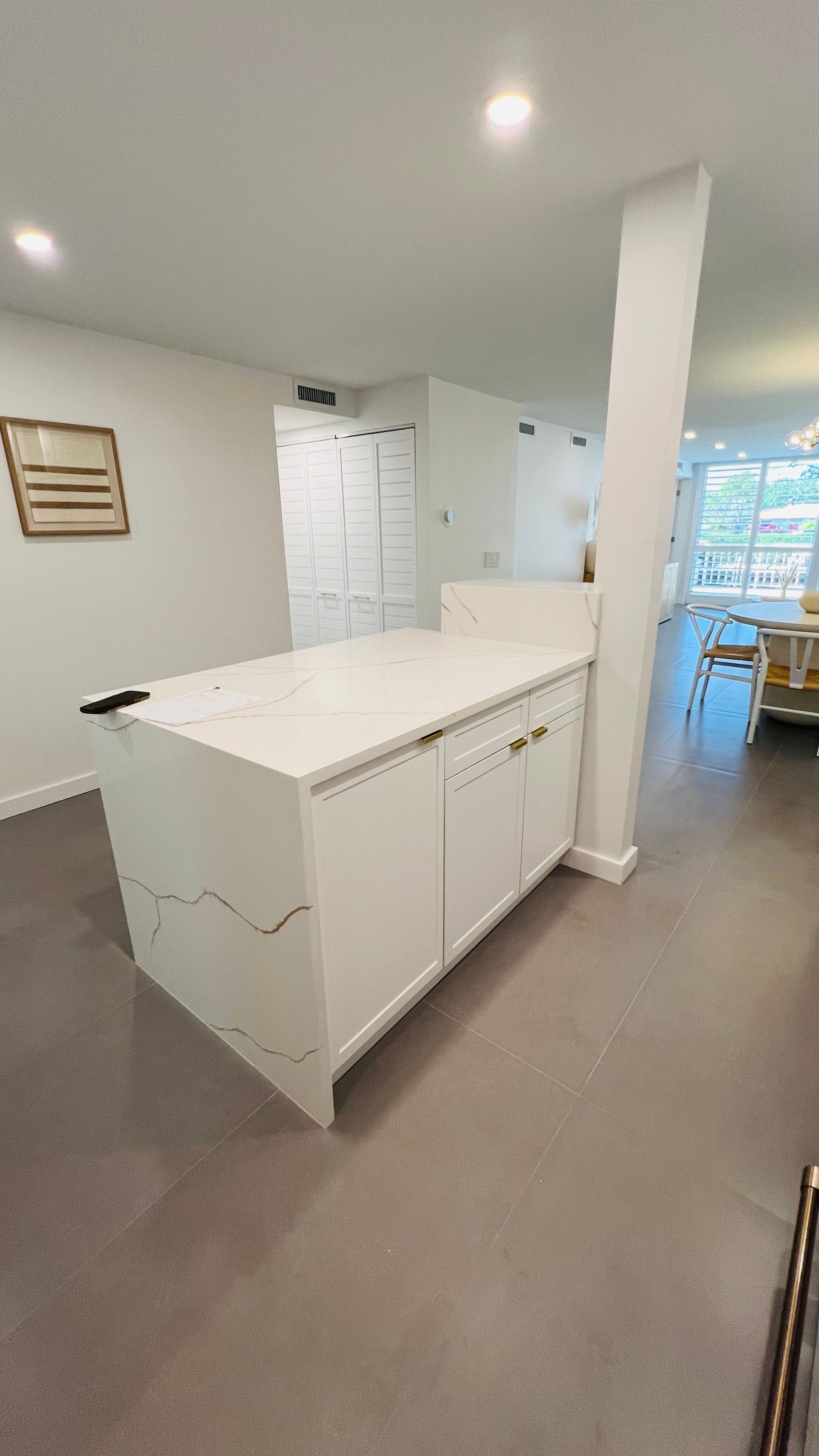 A white kitchen island with a marble countertop in a bright room with a support pillar and light wood flooring.