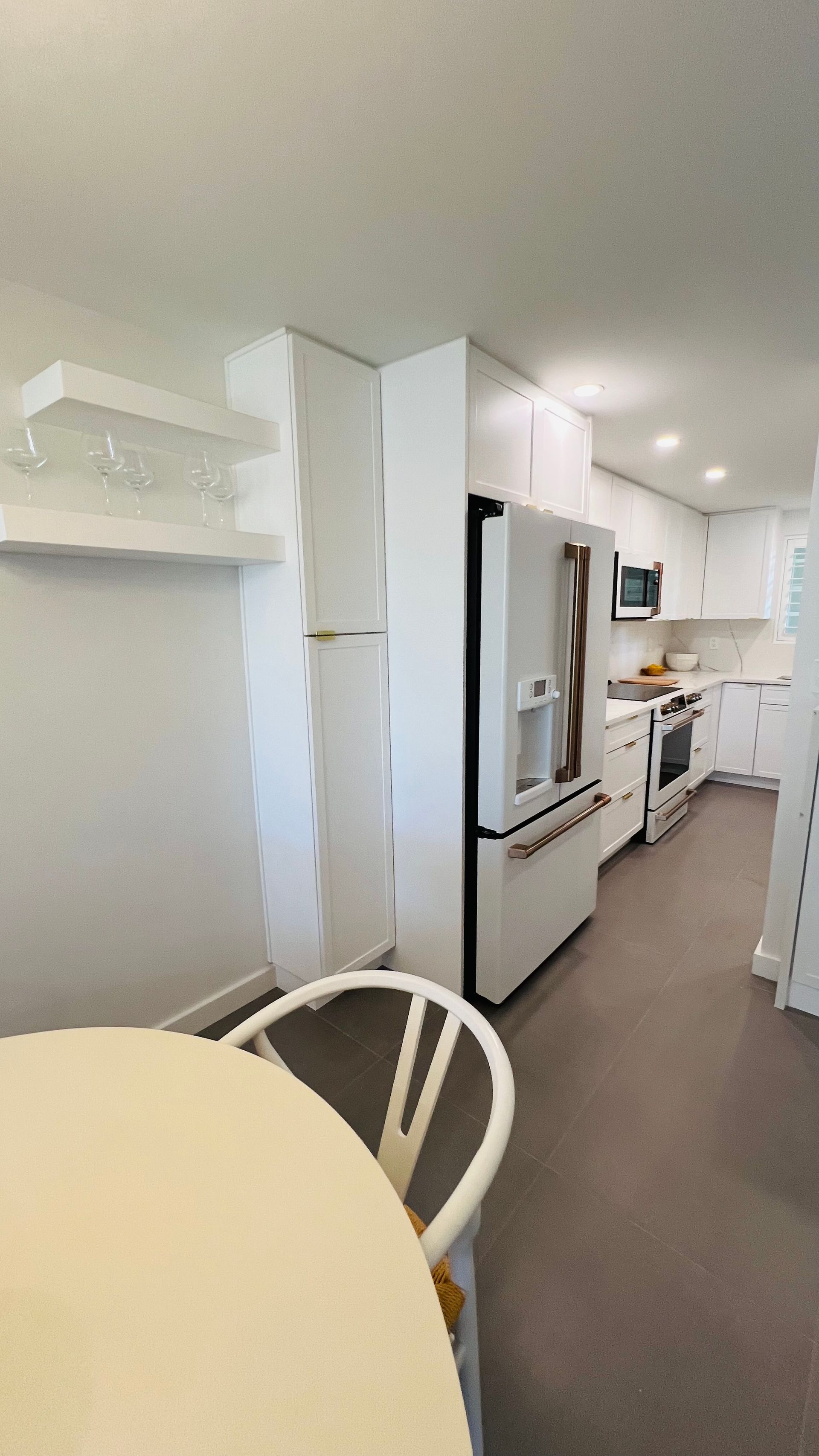 A white kitchen with a refrigerator, wall cabinets, floating shelves, and a portion of a dining table in the foreground.
