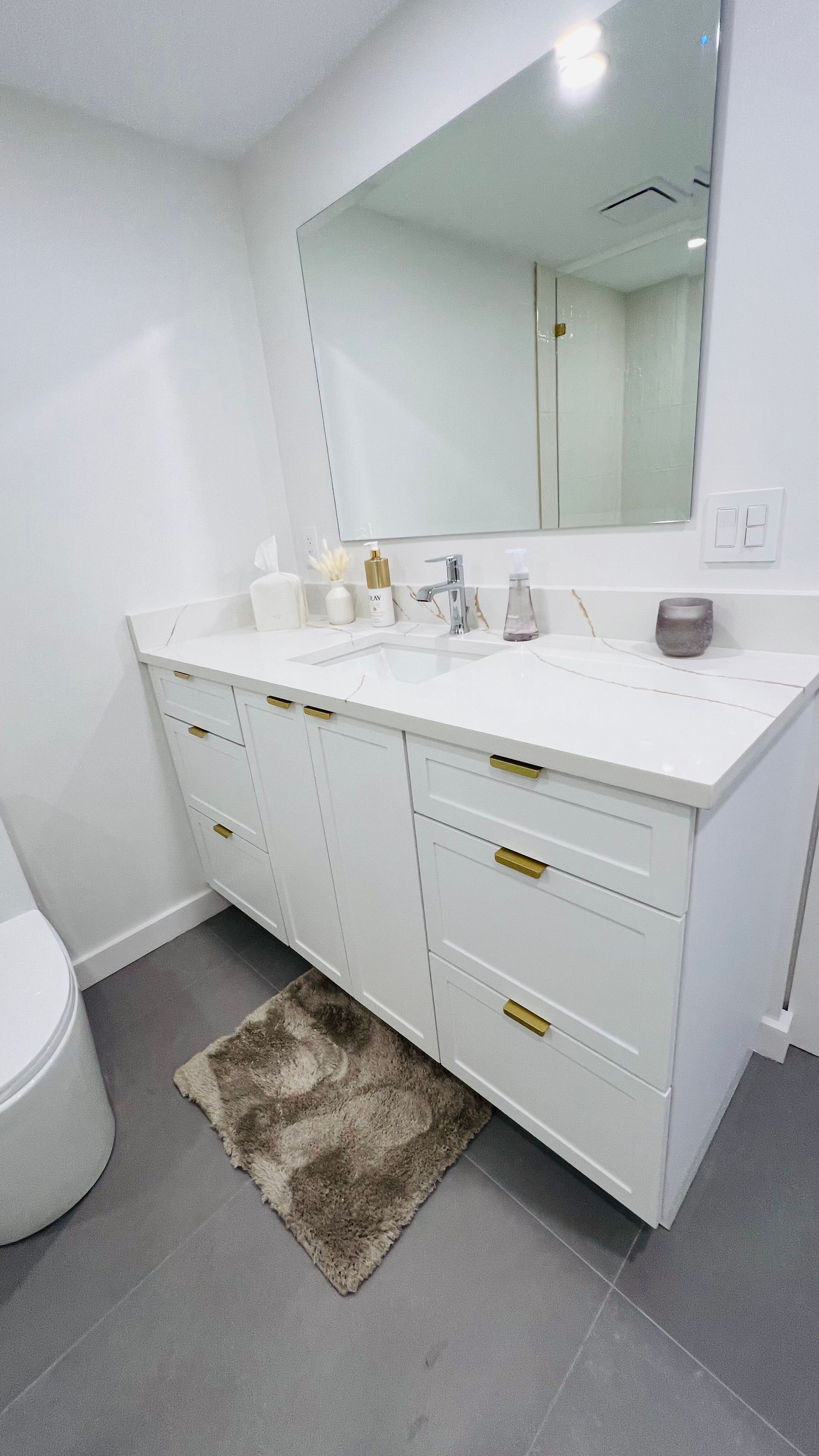 A modern bathroom vanity with a white countertop, gold hardware, and a large mirror against a white wall.