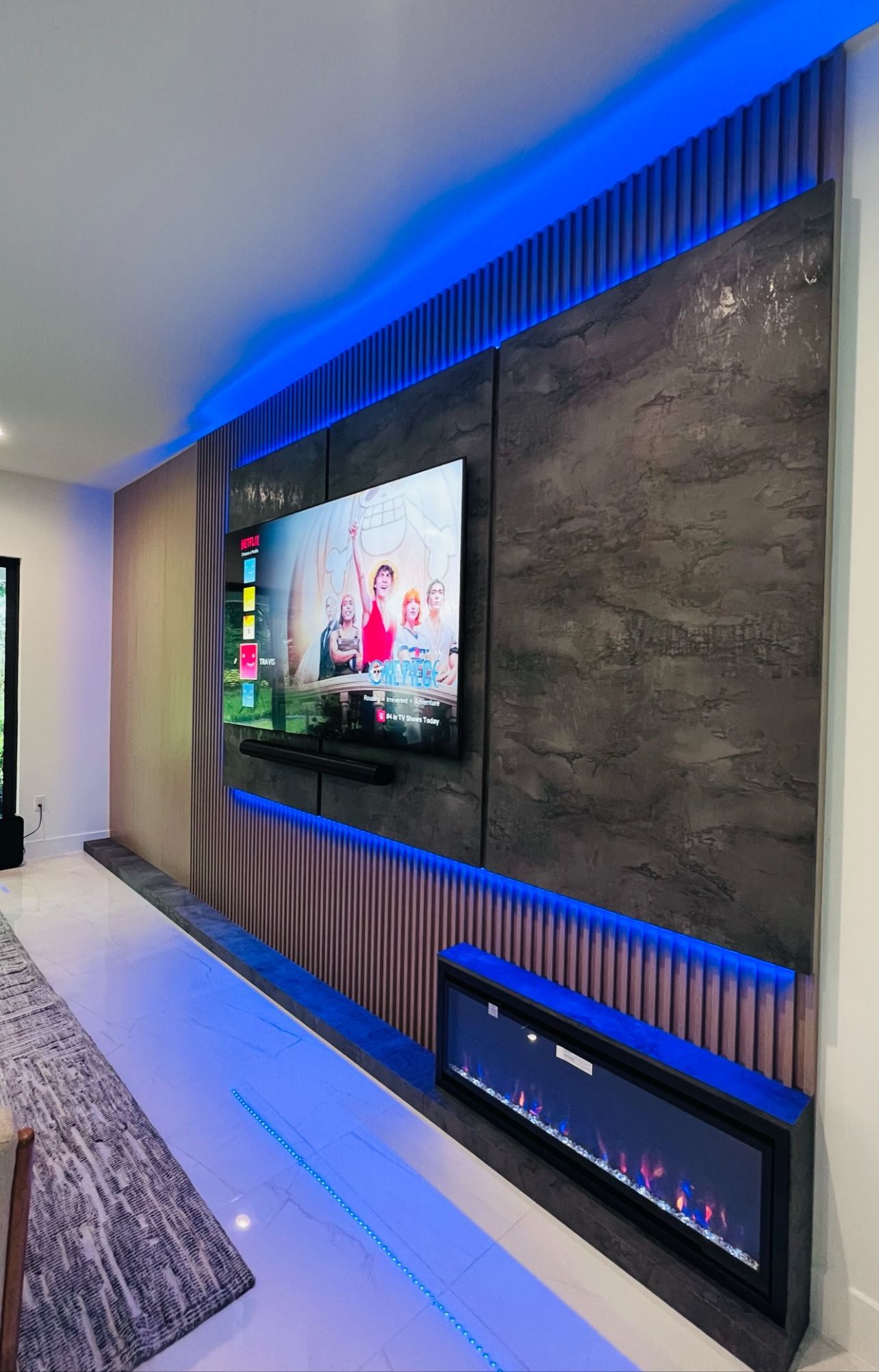A modern wall featuring a TV, fireplace, and textured dark panels with blue LED accent lighting.