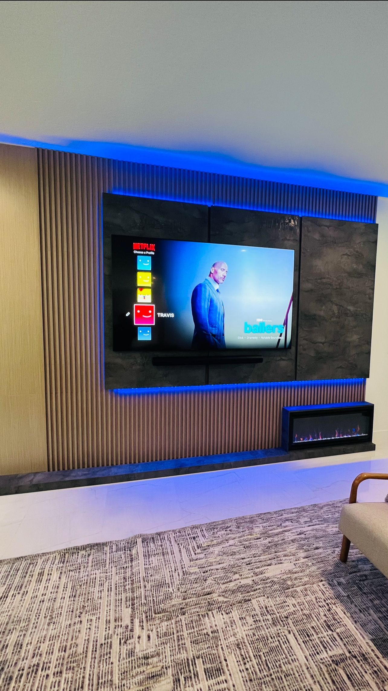 A mounted TV with blue LED accent lighting on a textured, wood-slat and stone wall above a fireplace in a modern room.