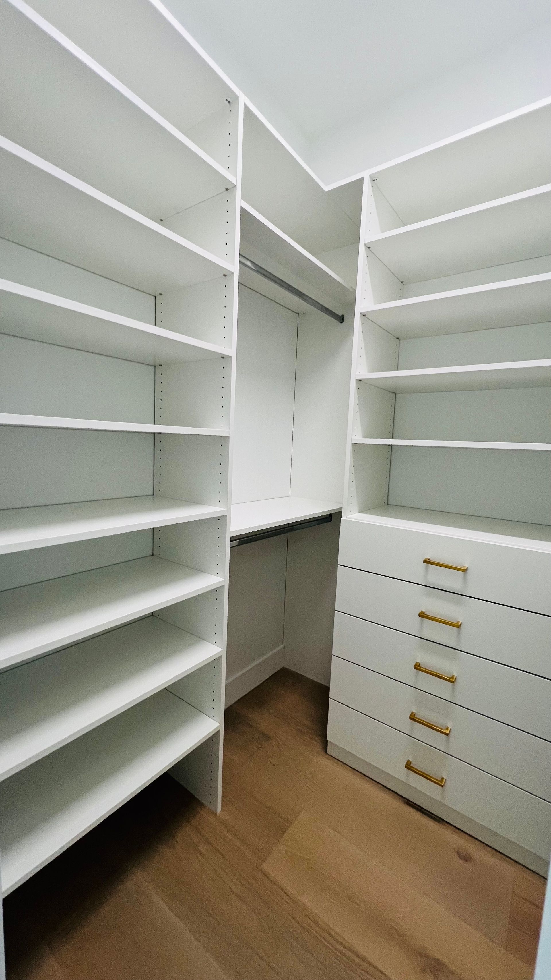 Walk-in closet with white shelving, a hanging rod, five drawers with gold handles, and light-toned wood flooring.