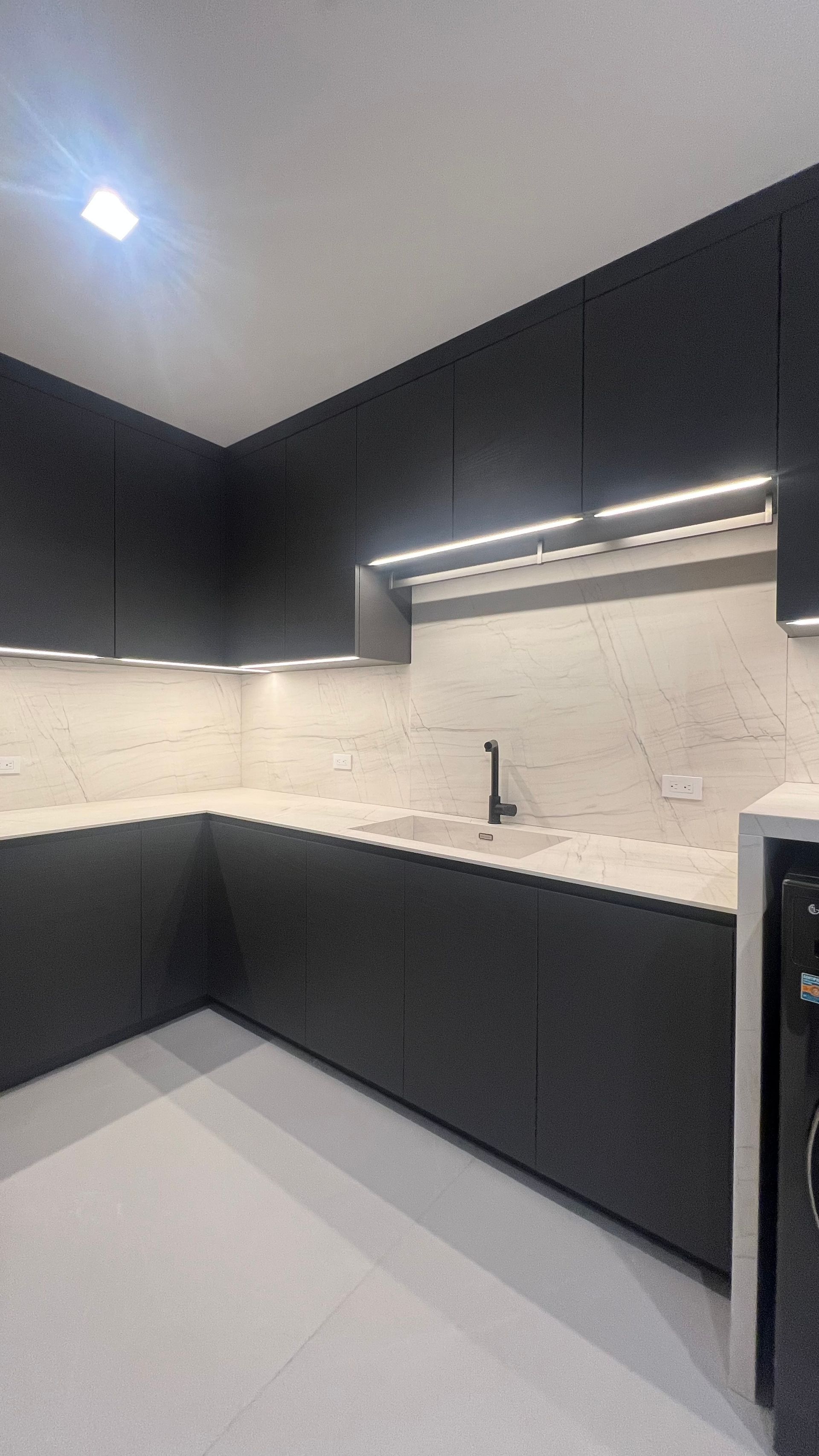 Modern L-shaped kitchen with dark matte cabinets, white stone countertops, under-cabinet lighting, and a single sink.