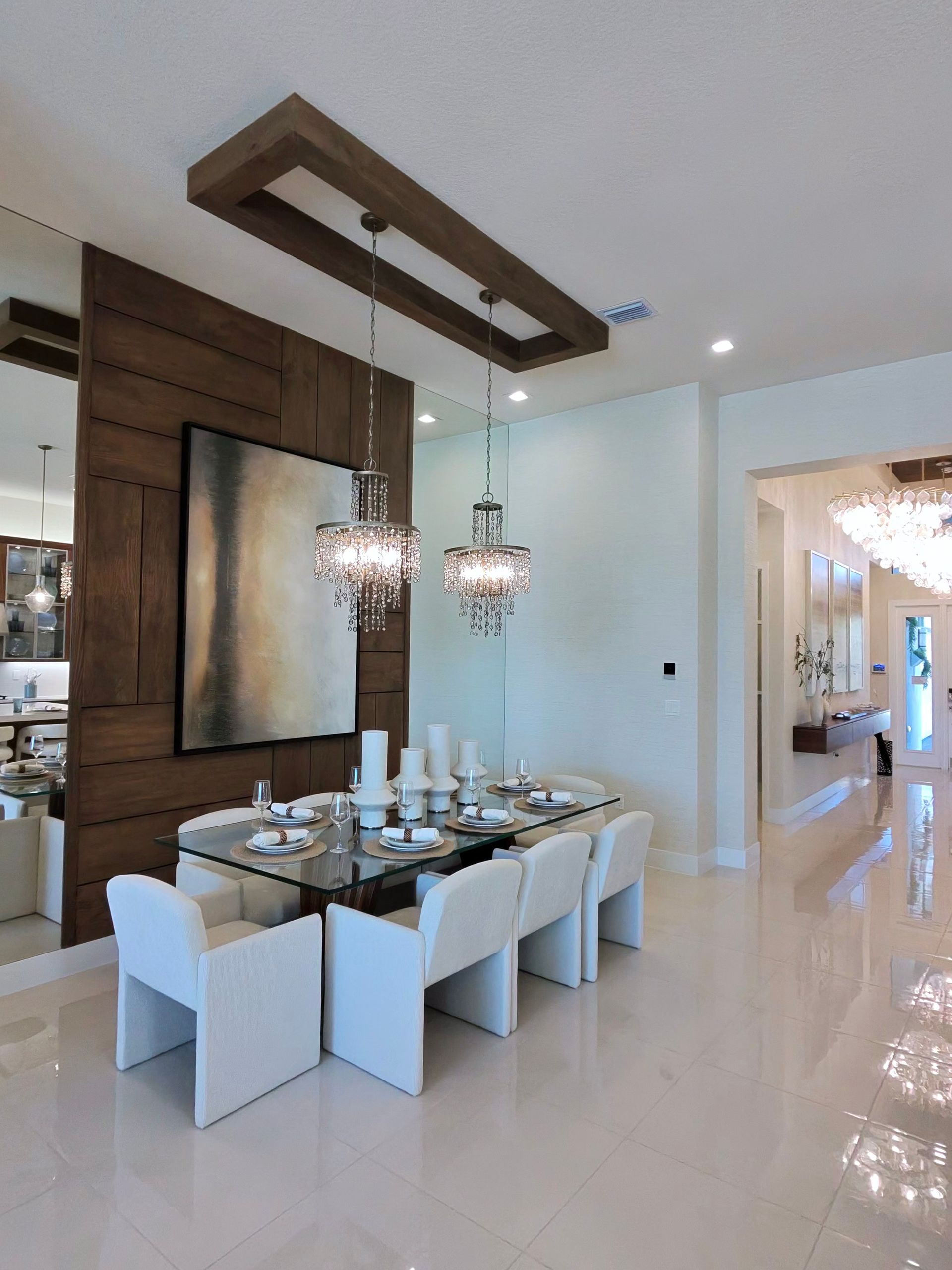 A modern dining room featuring a glass table with white chairs, hanging crystal lights, and a wood-paneled accent wall.