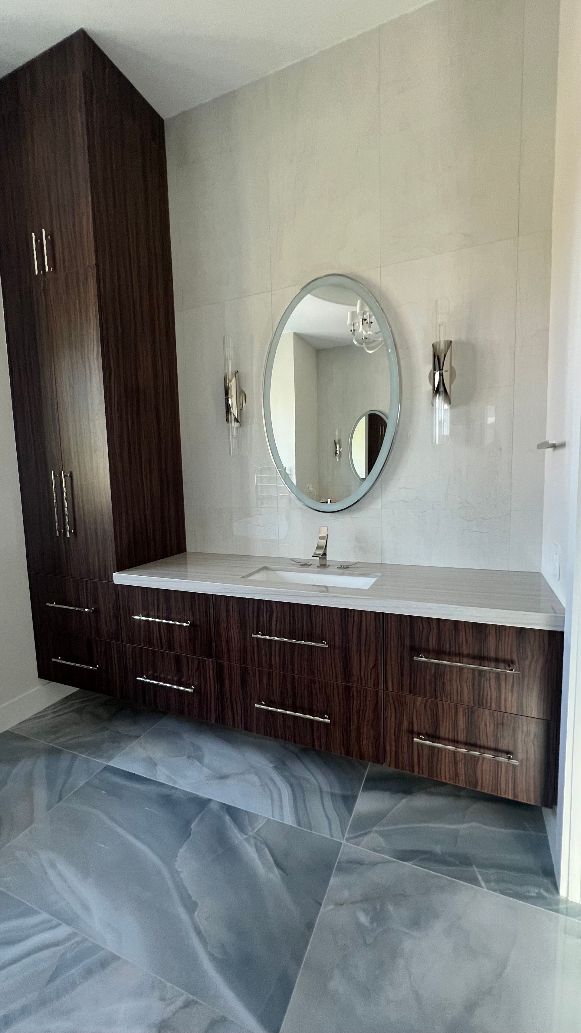 A modern bathroom featuring a wood-grain floating vanity, a tall matching cabinet, an oval mirror, and gray tiled floors.