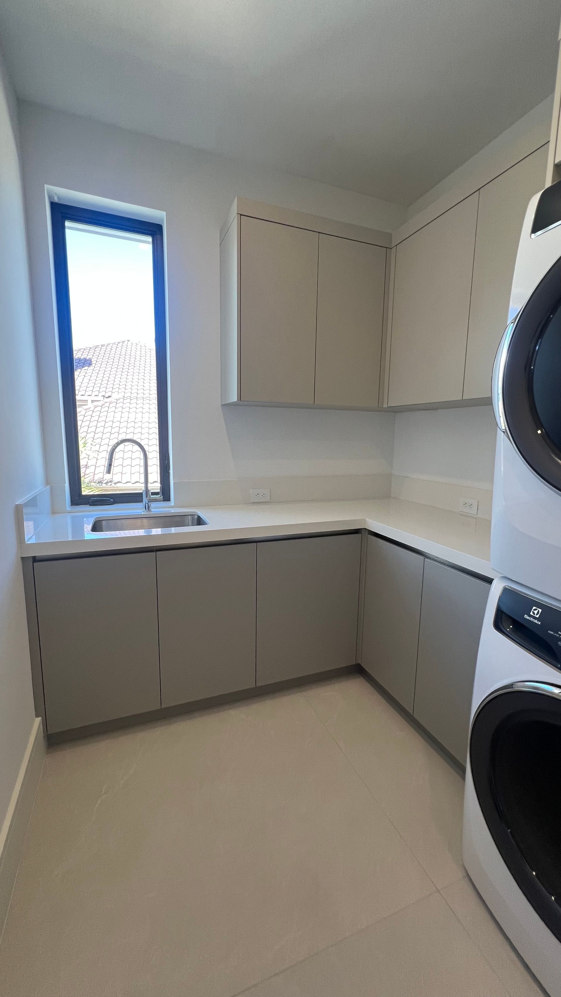 A laundry room featuring light gray cabinets, a white countertop with a stainless steel sink, and a stacked washer/dryer.