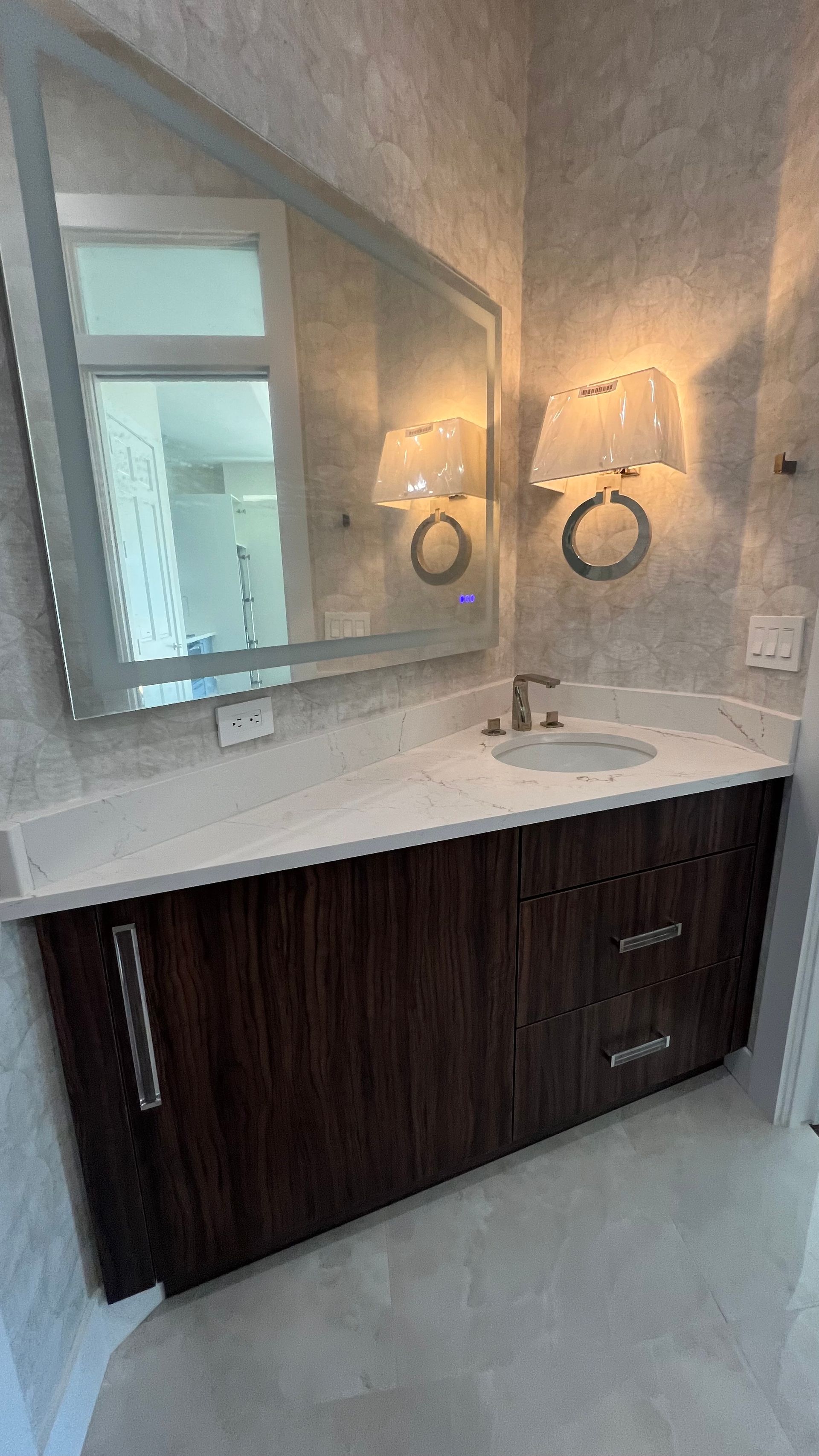 A bathroom vanity with a dark wood cabinet, white countertop, sink, mirror, and two wall sconces on a patterned wall.