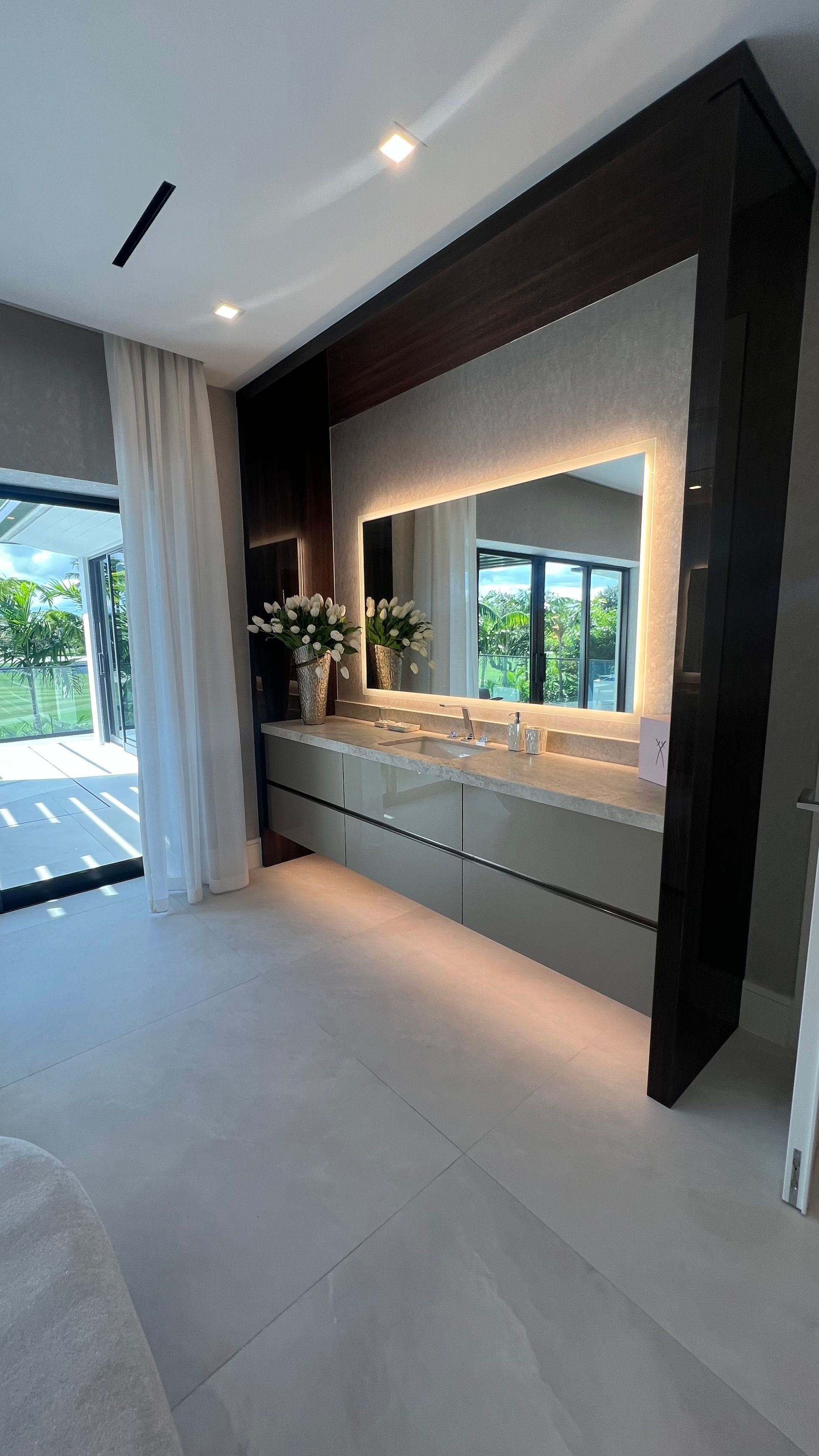 A modern bathroom vanity with a lighted mirror and dark wood surround, positioned next to a glass door with white curtains.