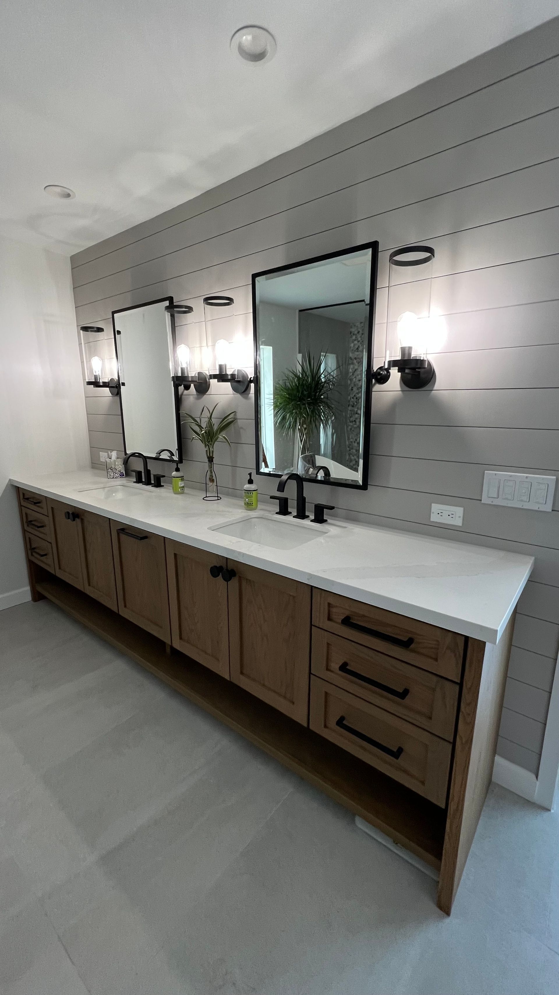 Double bathroom vanity with wood cabinets, white countertops, black fixtures, and mirrors on a gray shiplap wall.