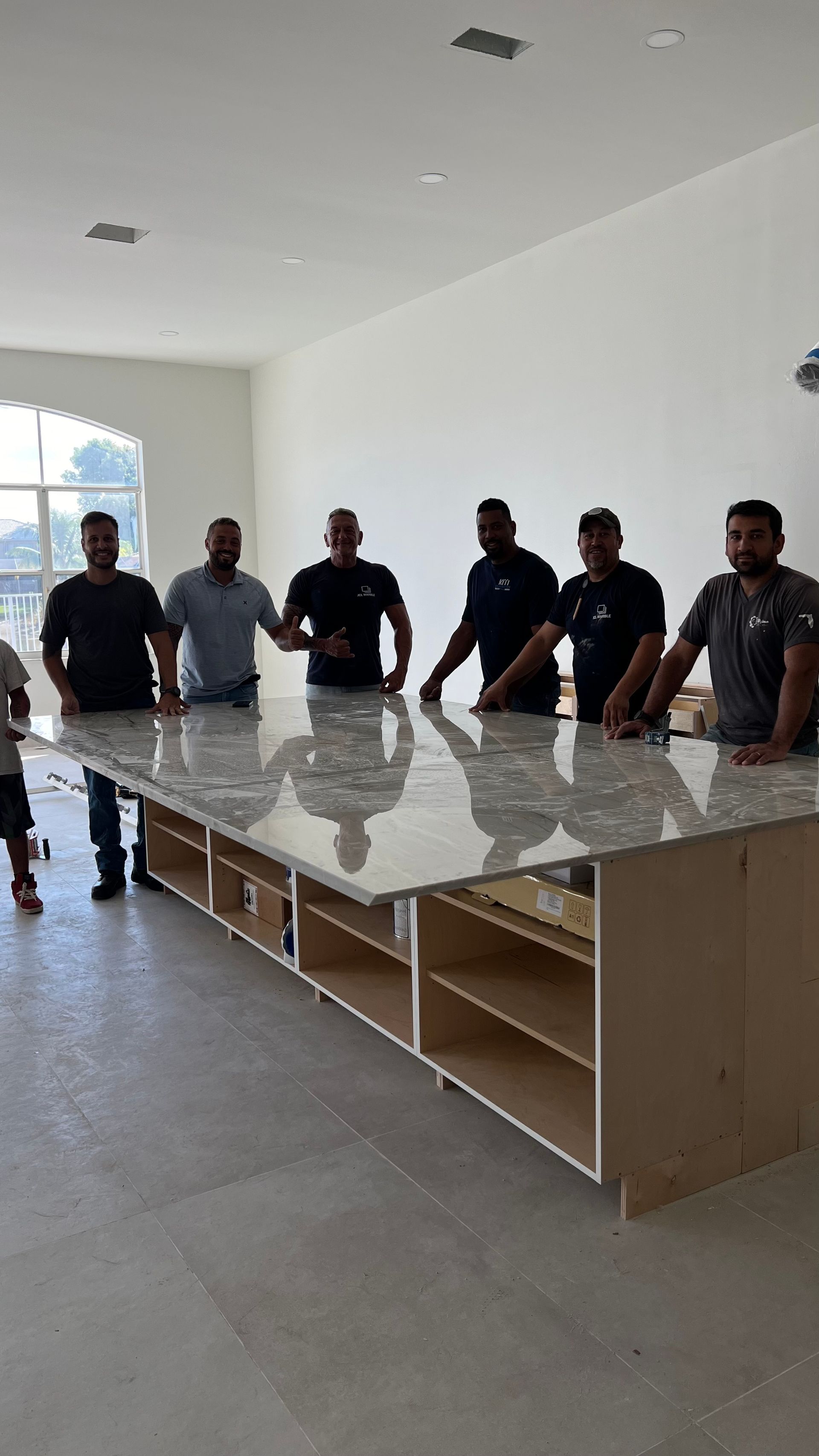 Seven people standing behind a long, unfinished kitchen island with a light granite countertop in a bright room.