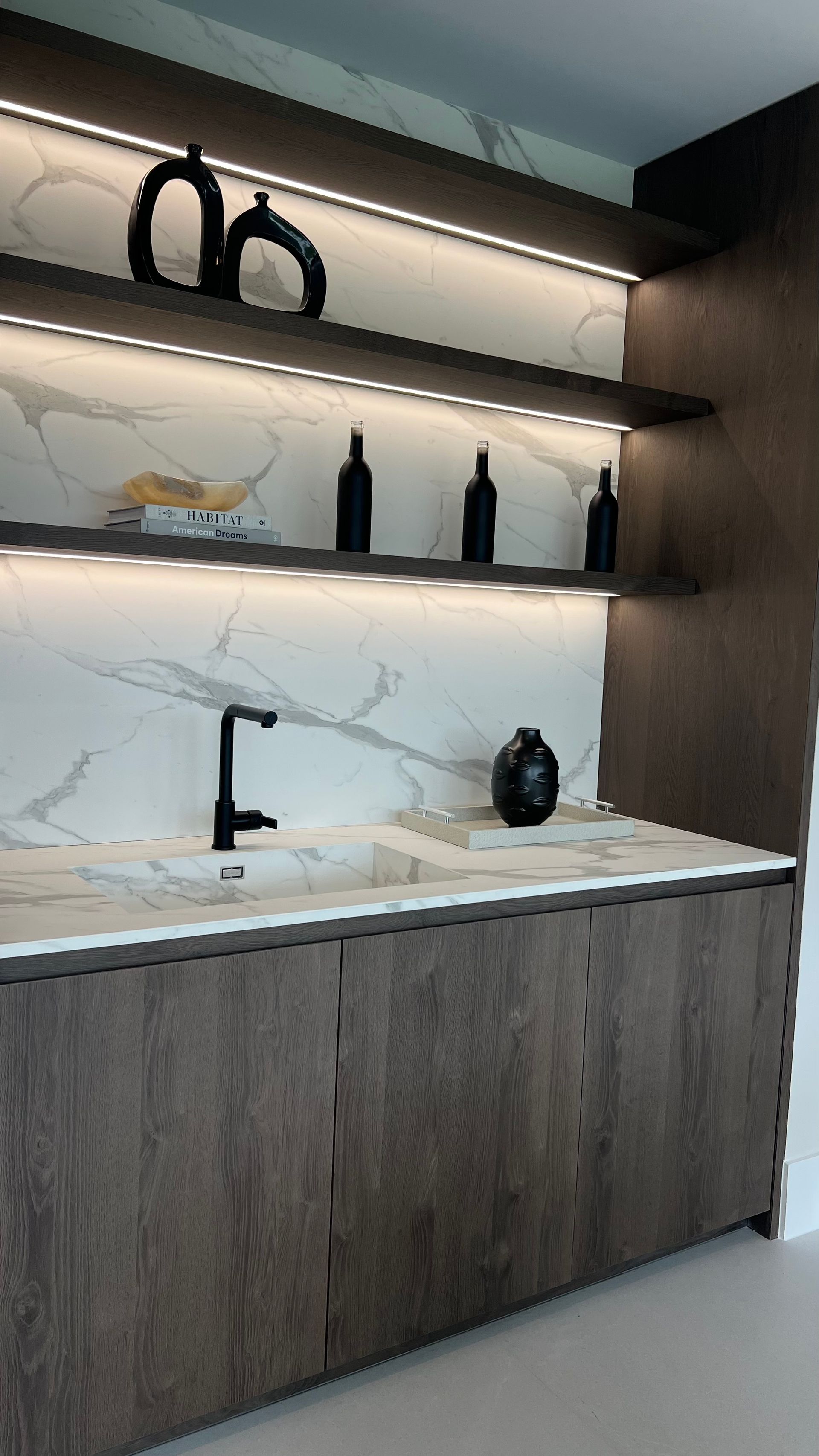 A modern wet bar with wooden cabinets, marble backsplash, floating shelves with LED lighting, and a black faucet.