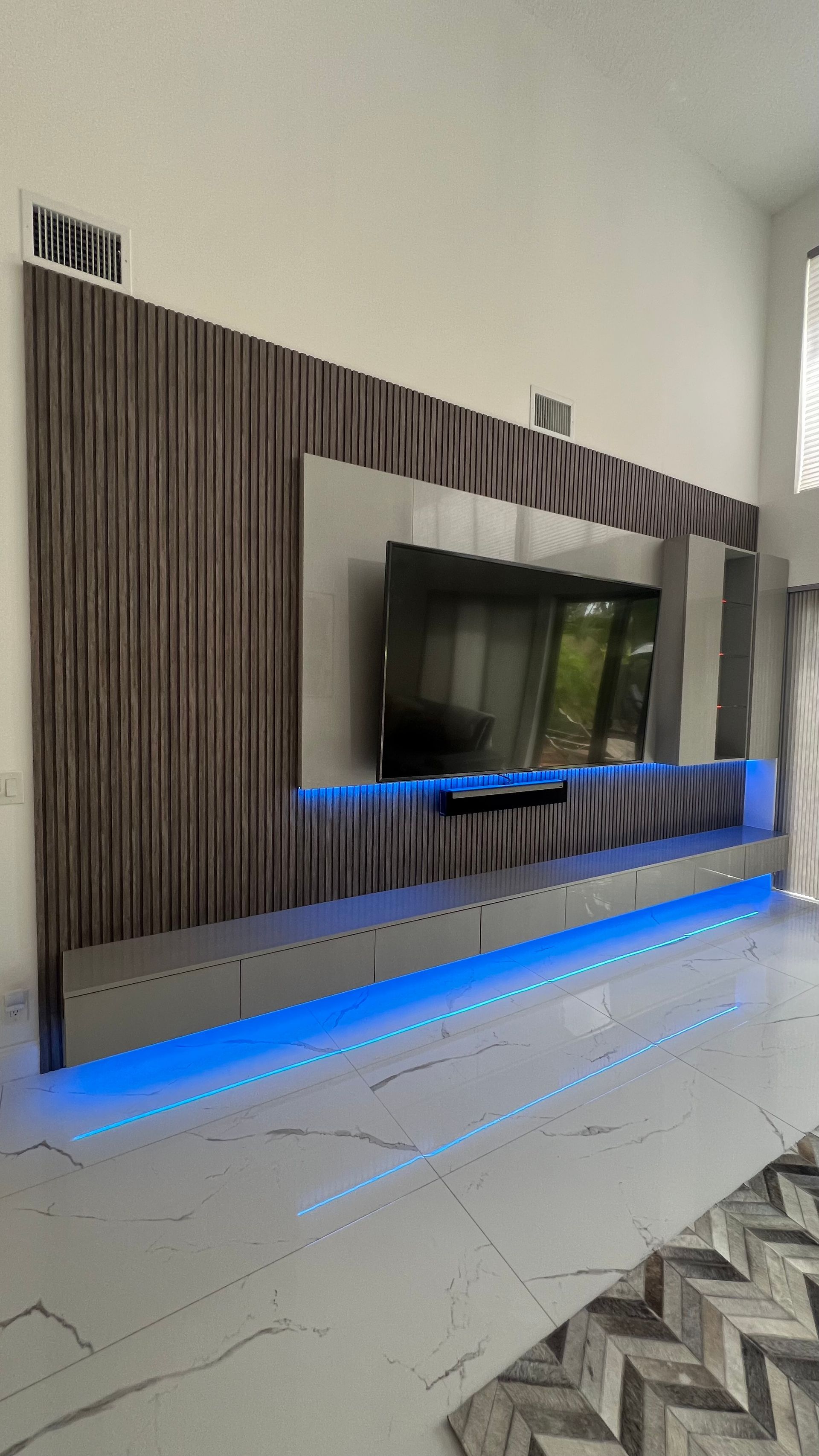 A wall-mounted TV unit with wood-slat panels, gray accents, and blue under-cabinet LED lighting in a modern room.