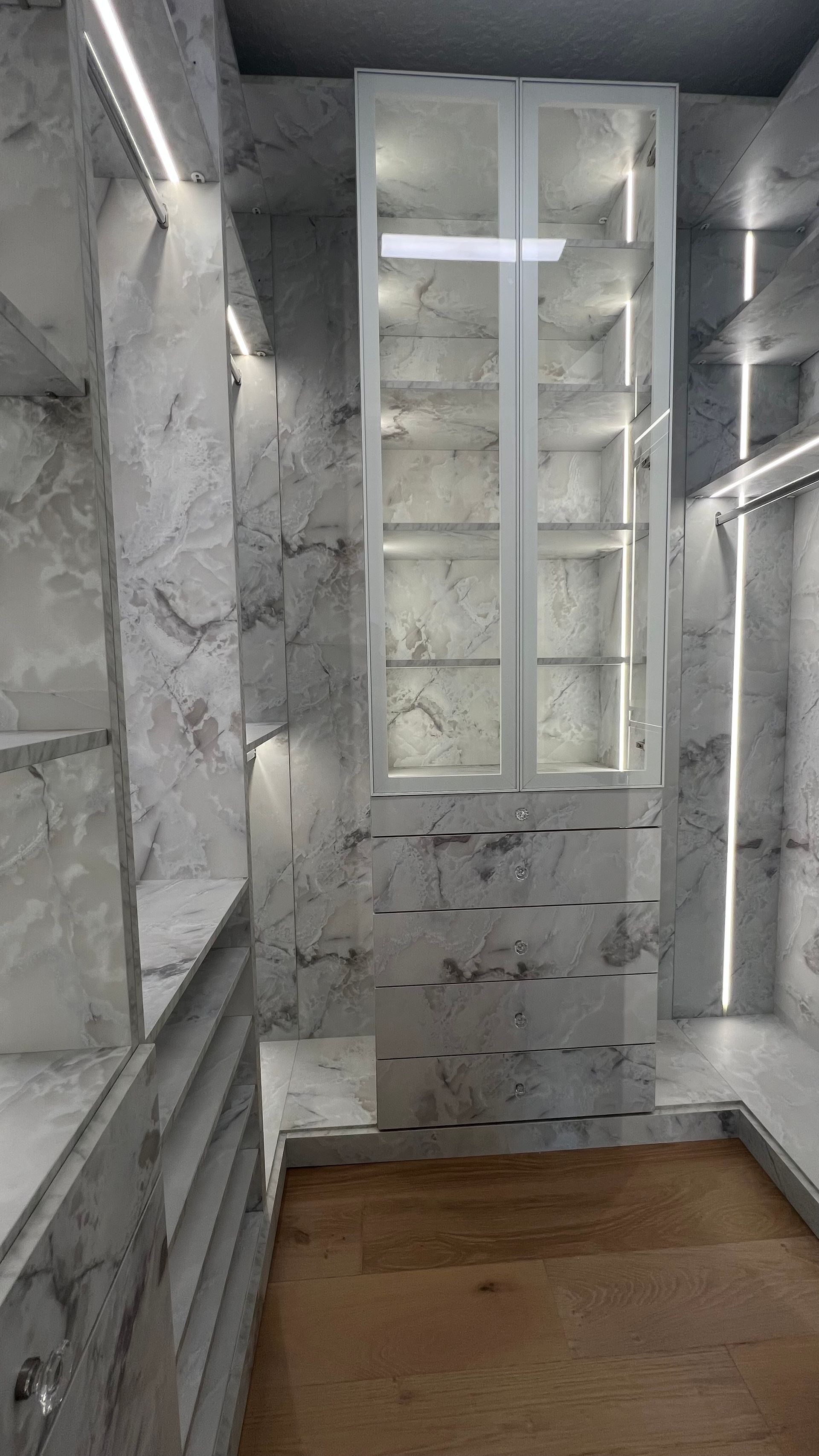 A modern walk-in closet featuring white marble-patterned cabinetry, integrated LED lighting, and light wood flooring.