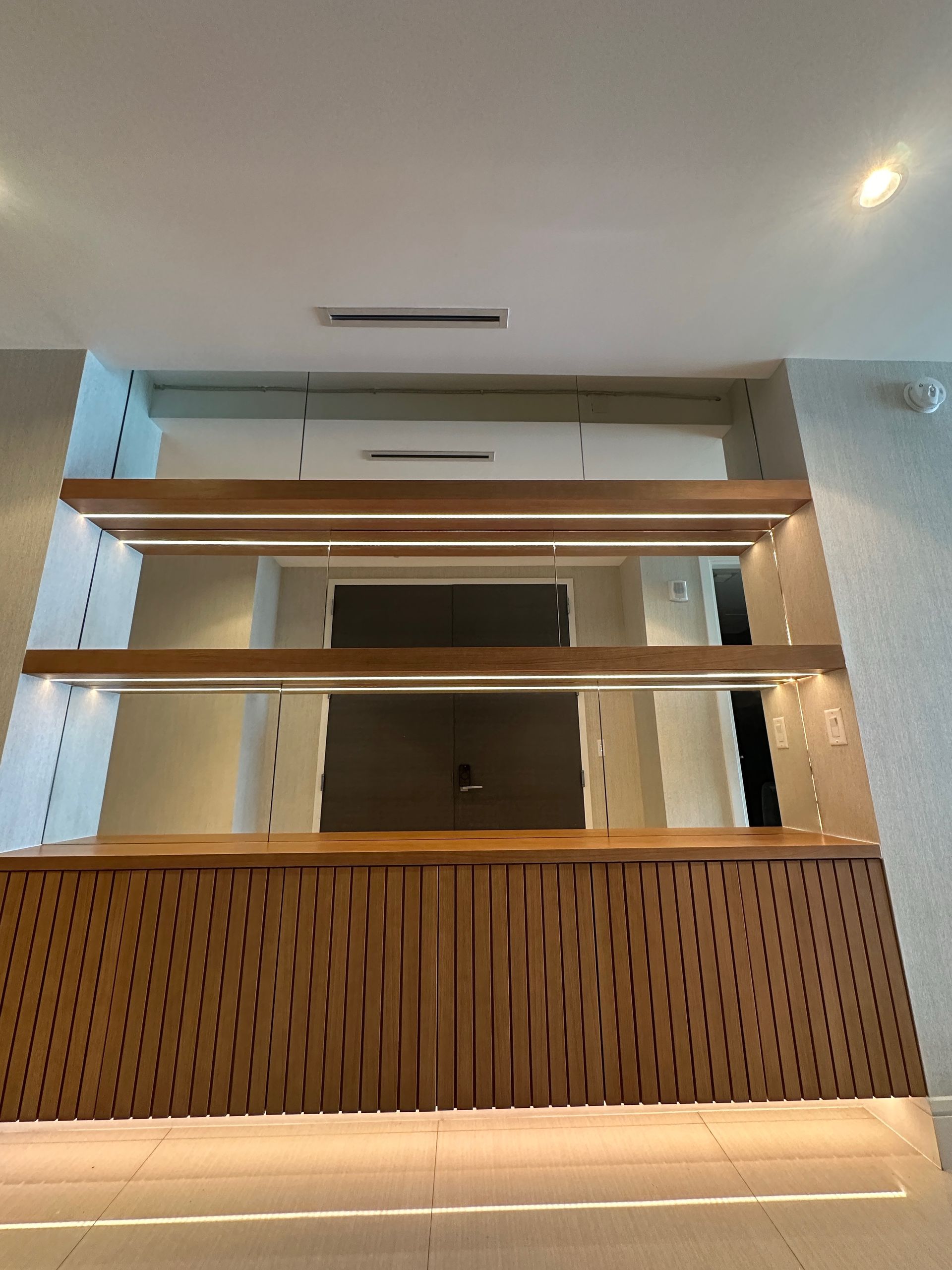 A modern built-in bar or display unit with a slatted wooden base, two floating shelves, and integrated lighting.