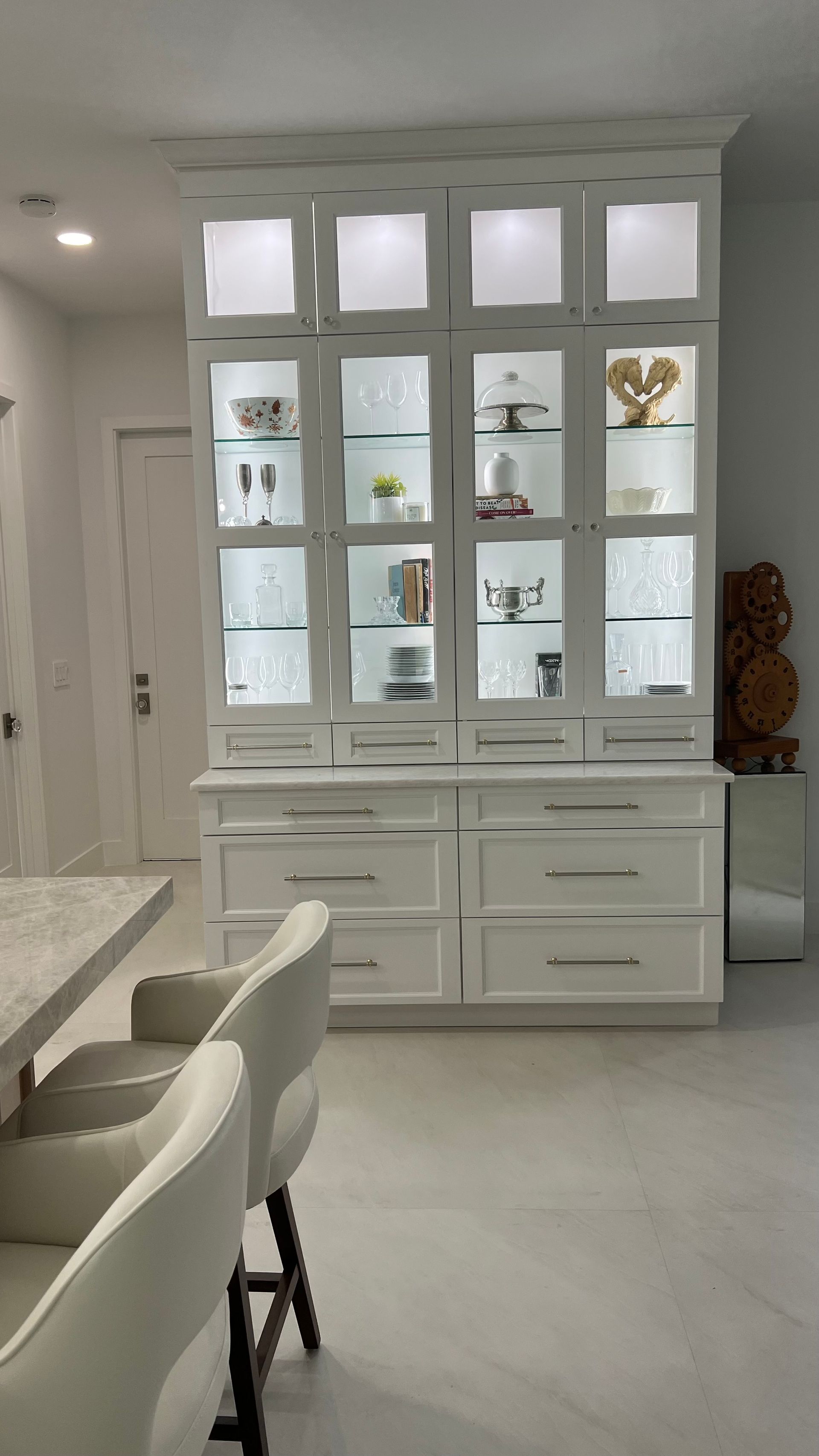 A large, white wooden china cabinet with glass doors, interior lighting, and lower drawers stands in a bright room.