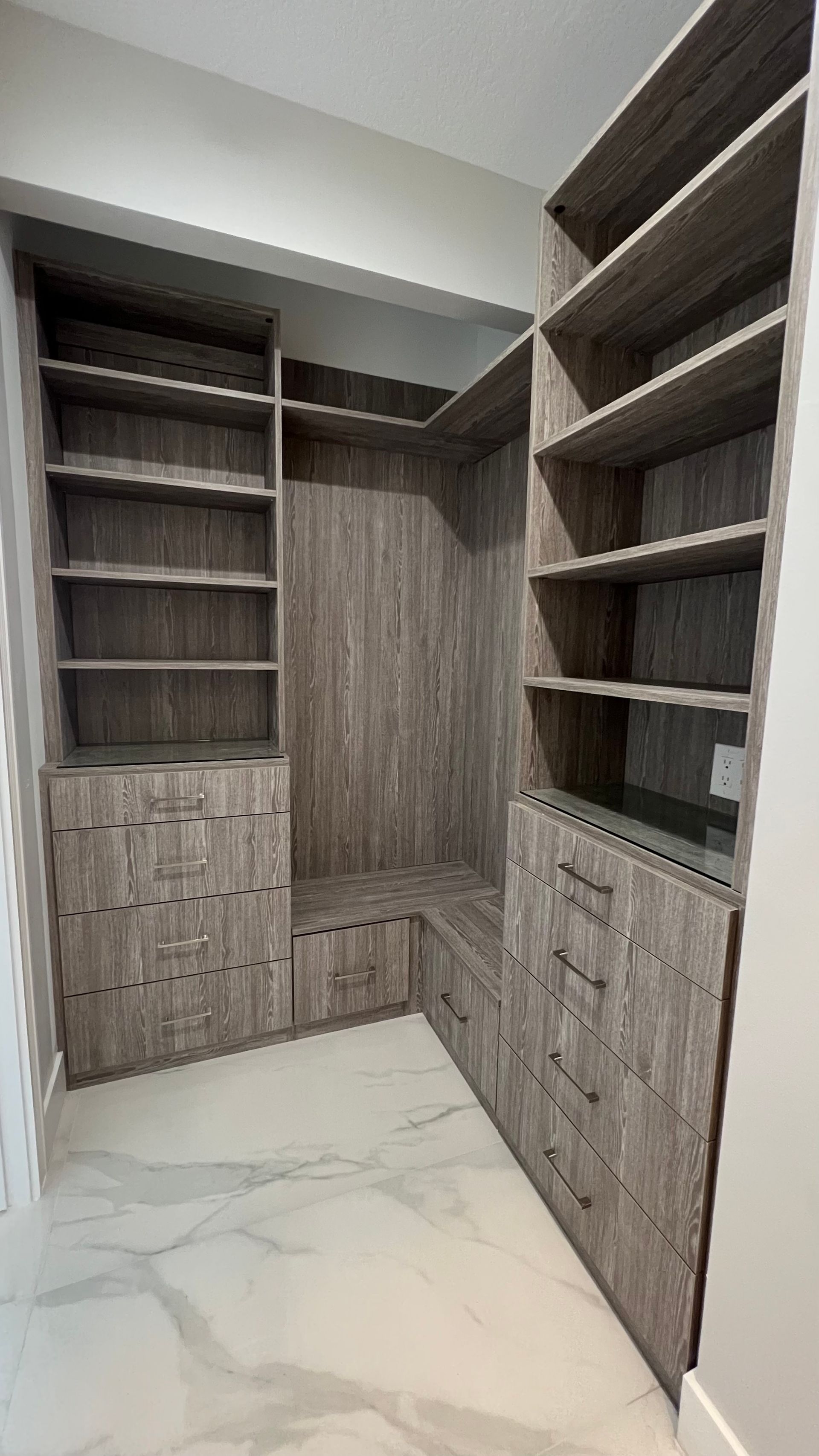 L-shaped wooden walk-in closet with open shelving and drawers, set against white marble-style flooring.