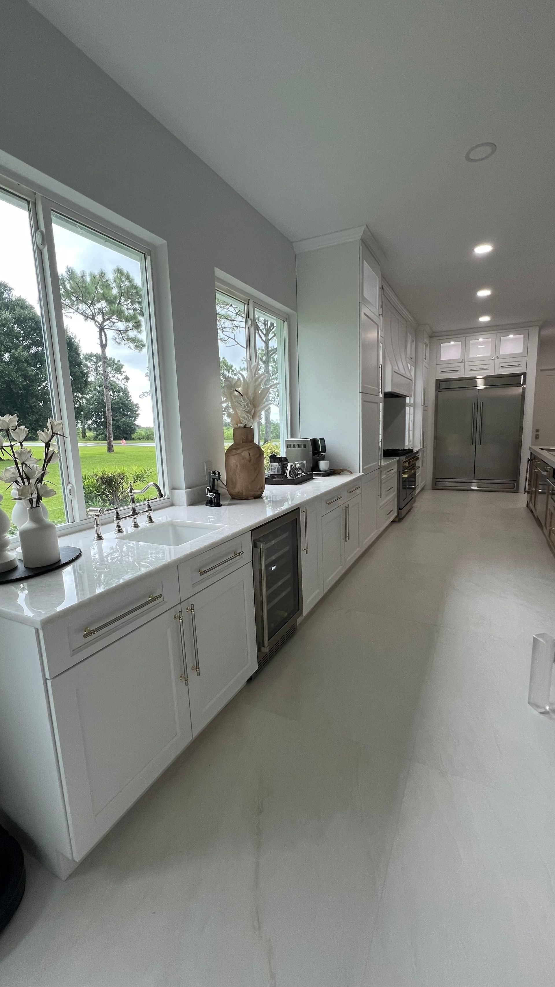 A bright, modern kitchen features long white counters, sleek white cabinetry, large windows, and light flooring.