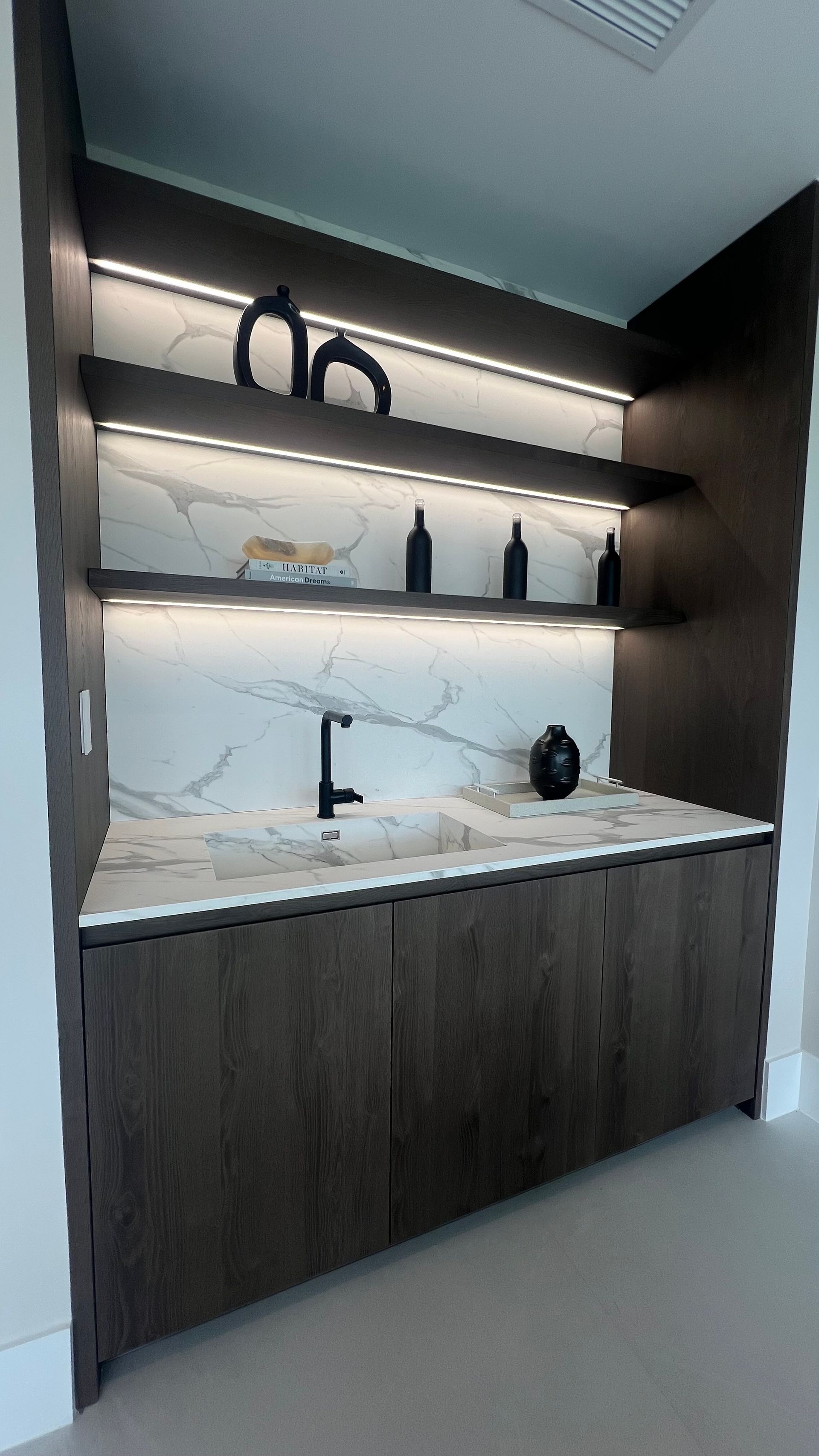 Modern dark wood wet bar with marble backsplash, white countertops, two illuminated shelves, and a black faucet.