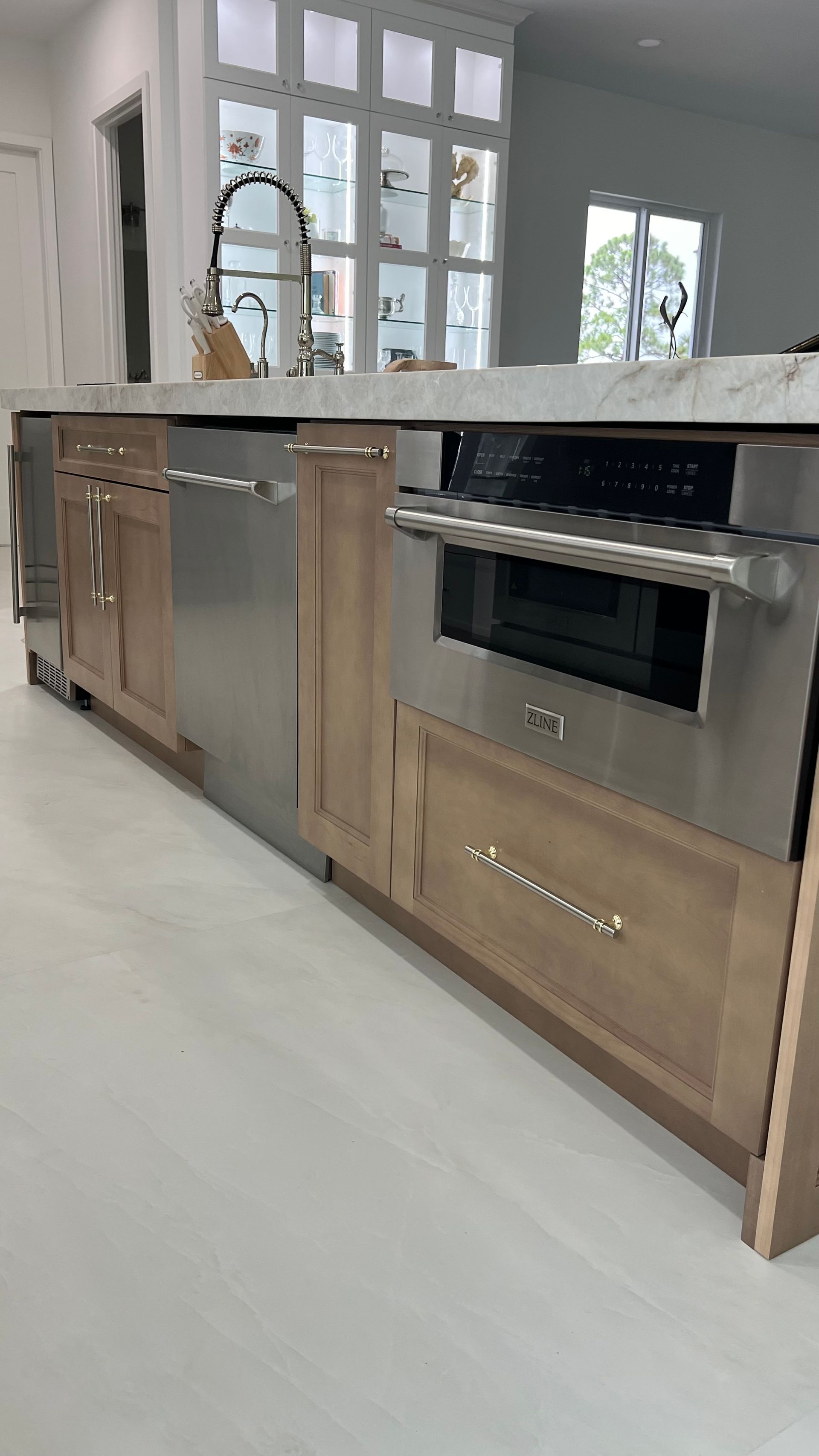 A kitchen island with natural wood cabinetry, a stainless steel dishwasher, a built-in microwave, and a white marble top.
