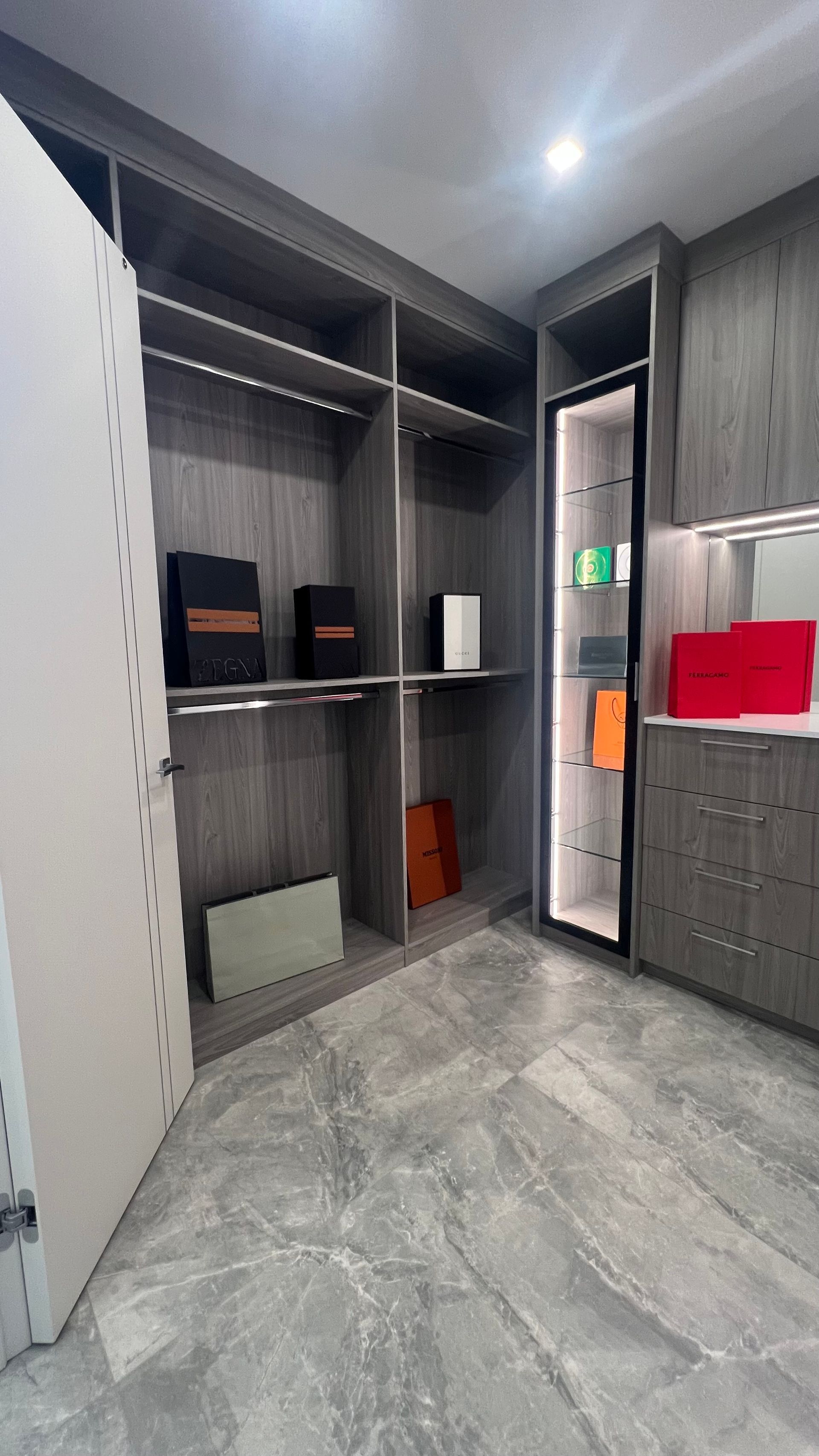 A walk-in closet featuring modern grey wood cabinetry, built-in shelving, a glass-front display case, and marble flooring.