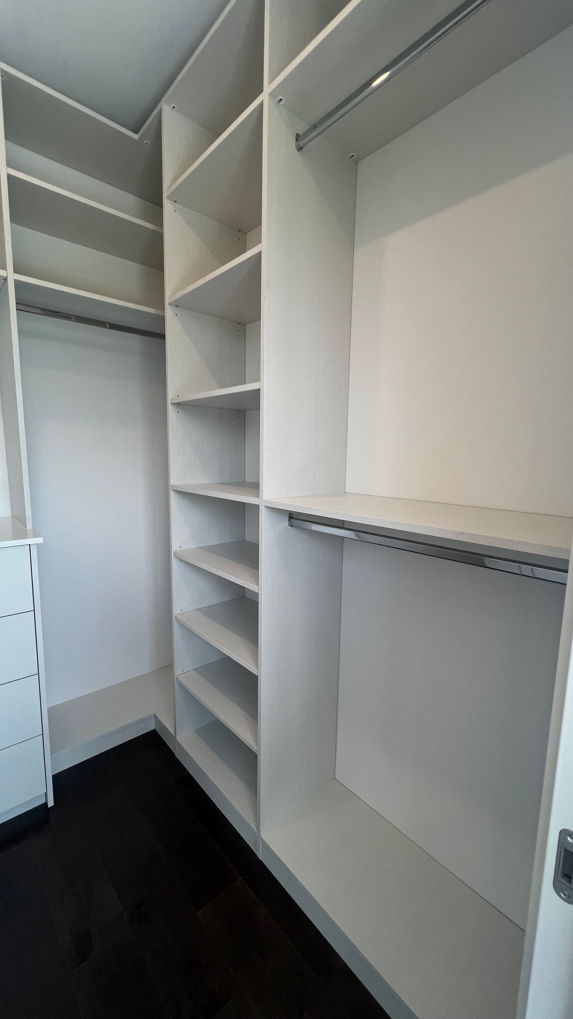 A walk-in closet featuring white wooden shelving units, hanging rods, and a set of drawers against a dark floor.