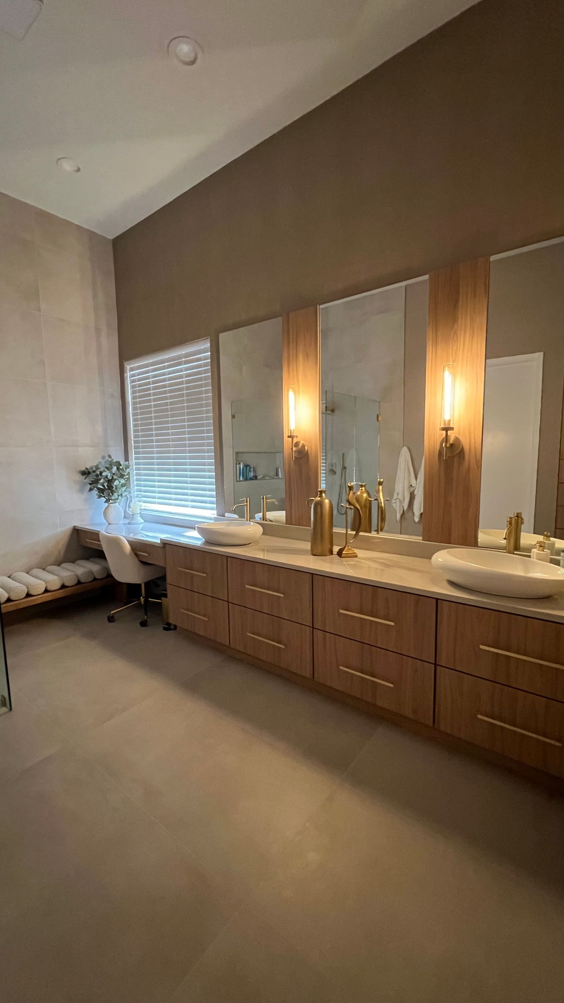 Modern bathroom with a long wooden vanity, double white sinks, large mirrors, warm lighting, and a window seat.