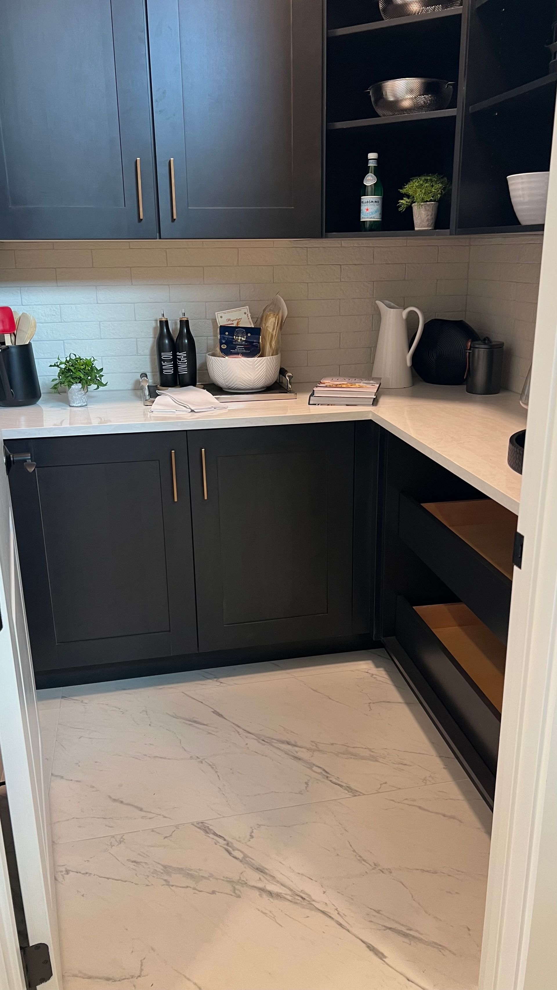 A walk-in pantry with dark cabinets, white quartz countertops, stone-look walls, and light-colored tile flooring.