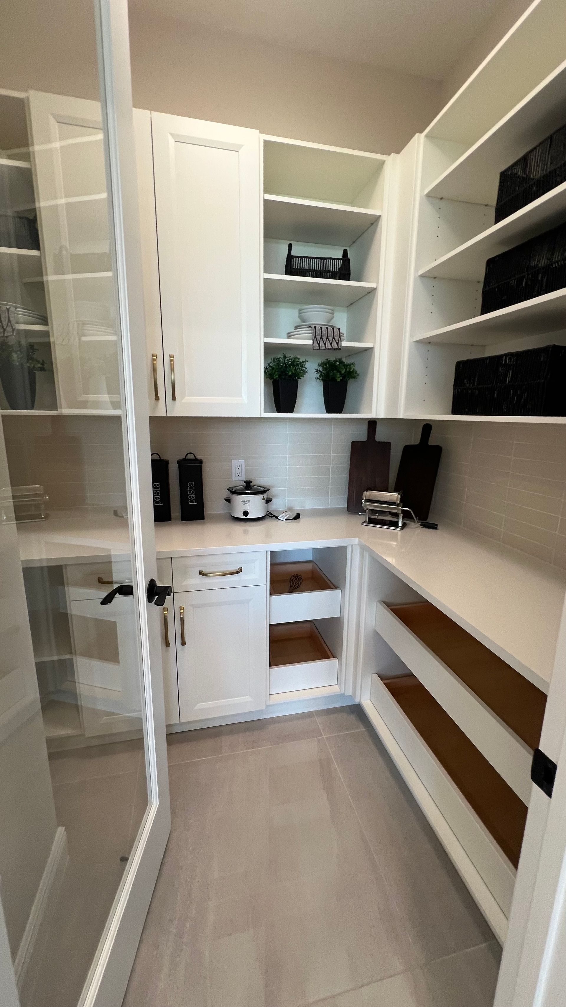A walk-in pantry with white cabinetry, open shelving, quartz countertops, a glass-paned door, and light tile flooring.