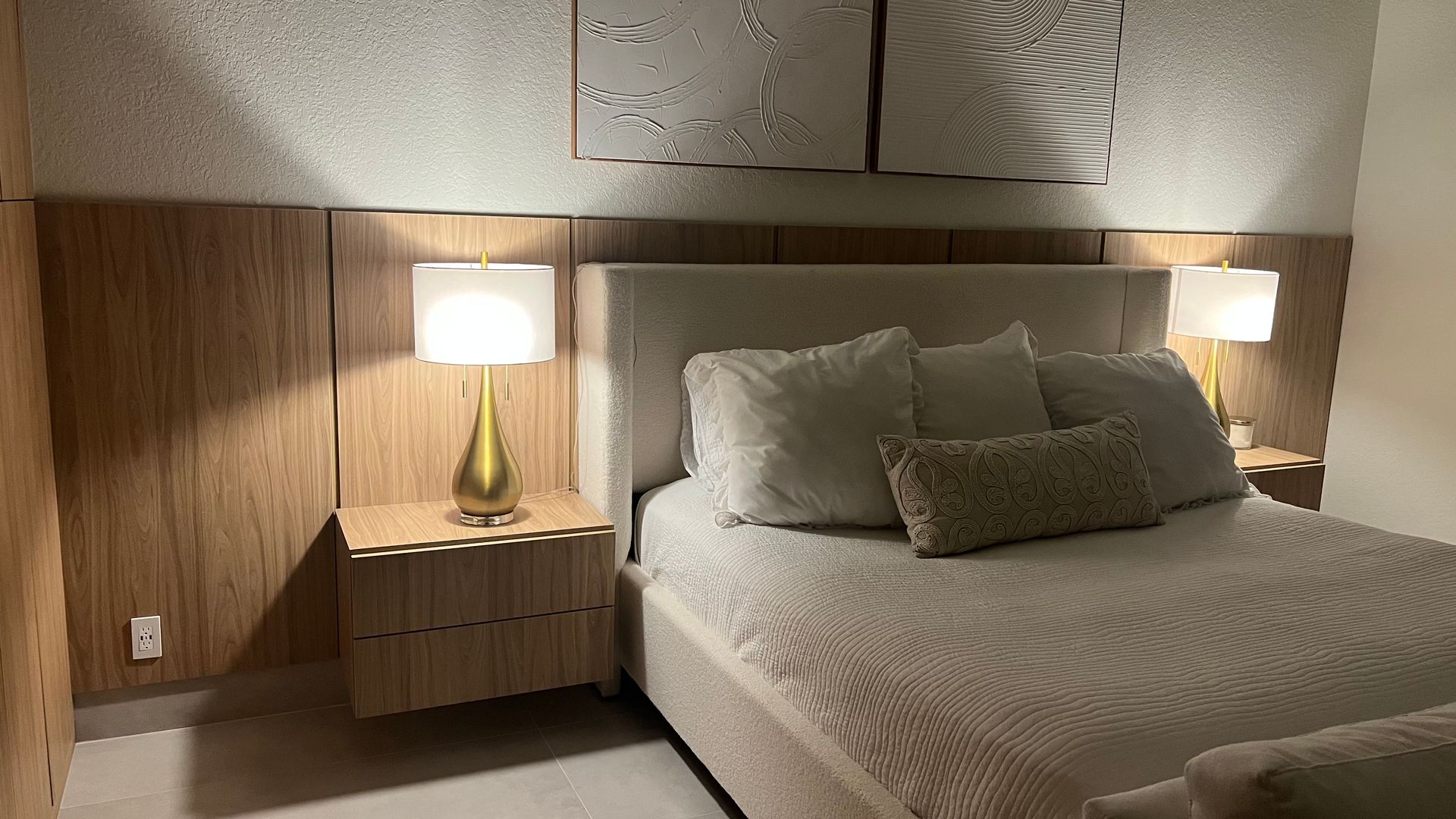 A bedroom with a bed featuring a cream headboard, a patterned bolster pillow, and two matching lamps on wood nightstands.