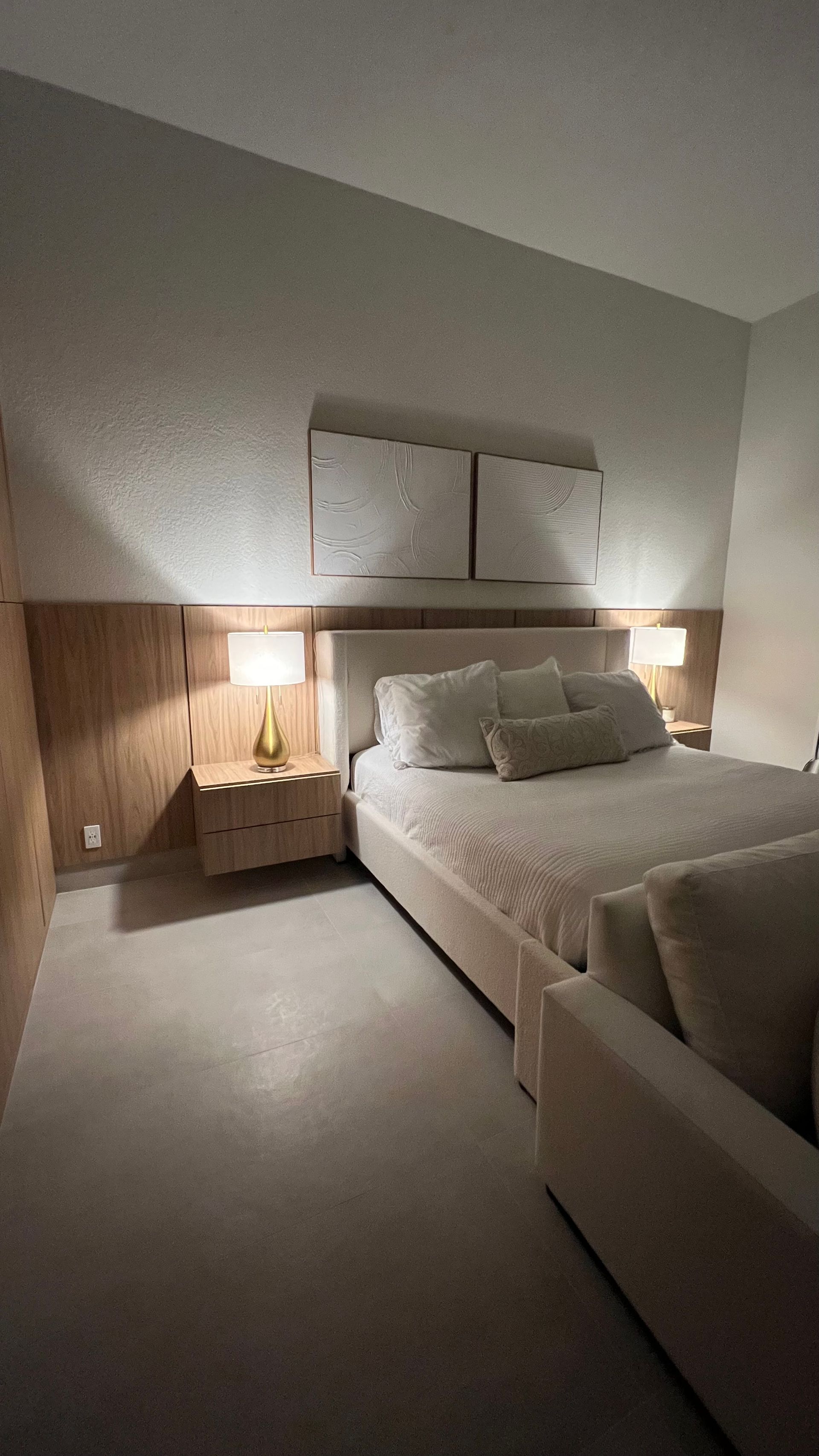 A bedroom featuring a bed with white bedding, wood-paneled wall, two bedside tables with lamps, and two framed wall artworks.