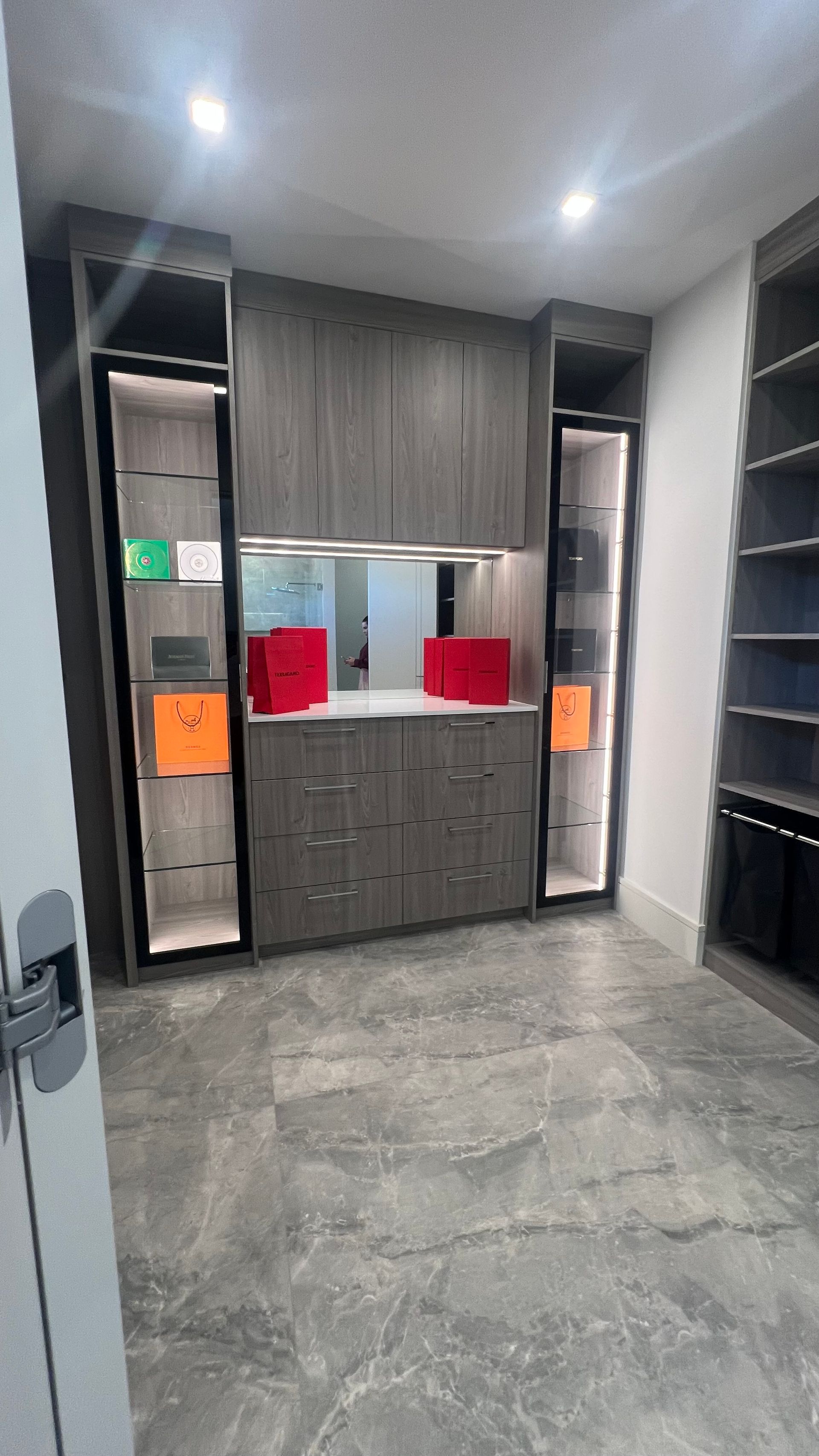 A custom walk-in closet featuring grey cabinetry, drawers, backlit glass displays with red boxes, and marble tile floors.