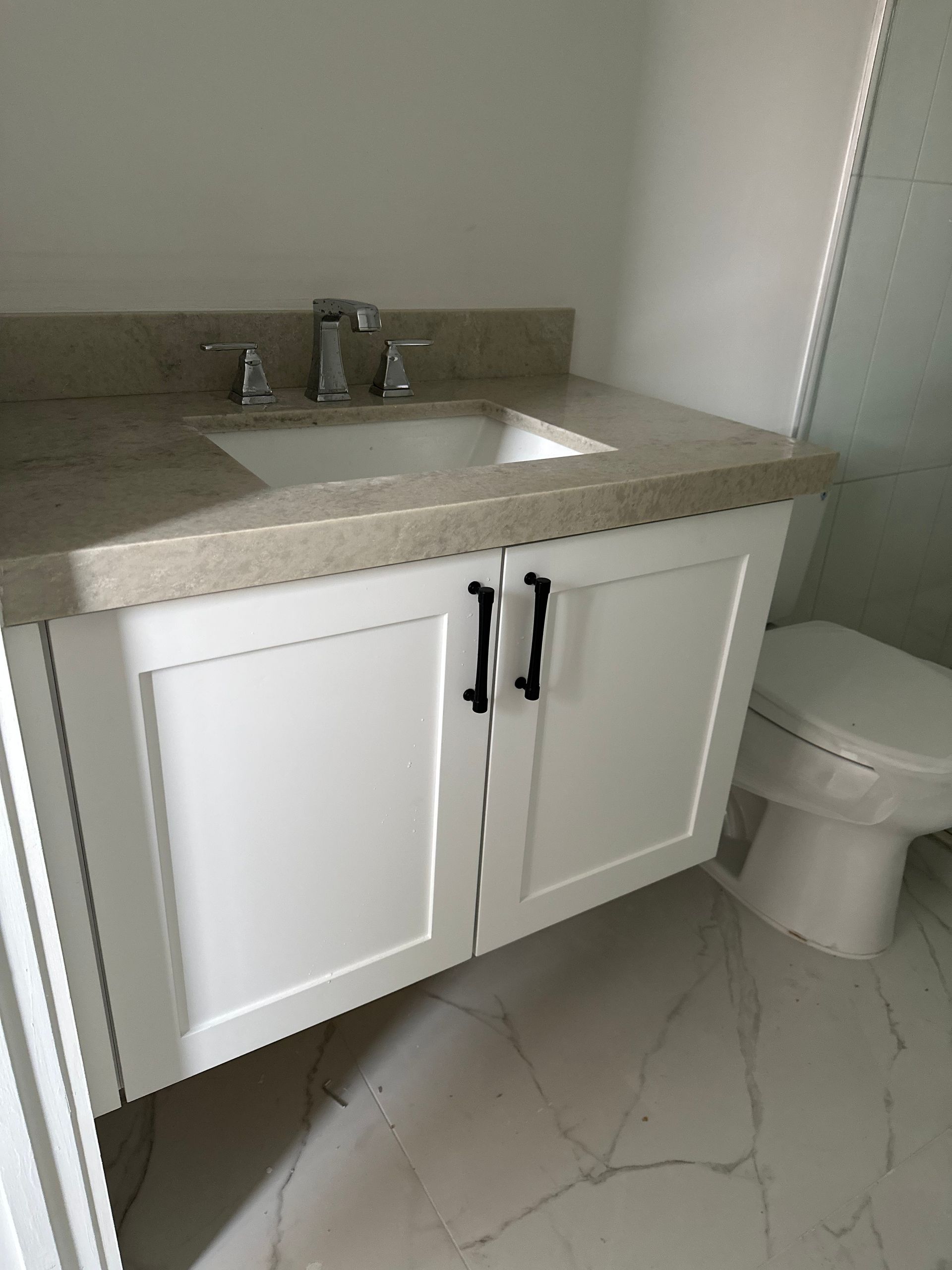 A white bathroom vanity with black hardware, a stone countertop, and a modern sink, next to a toilet on a marble floor.