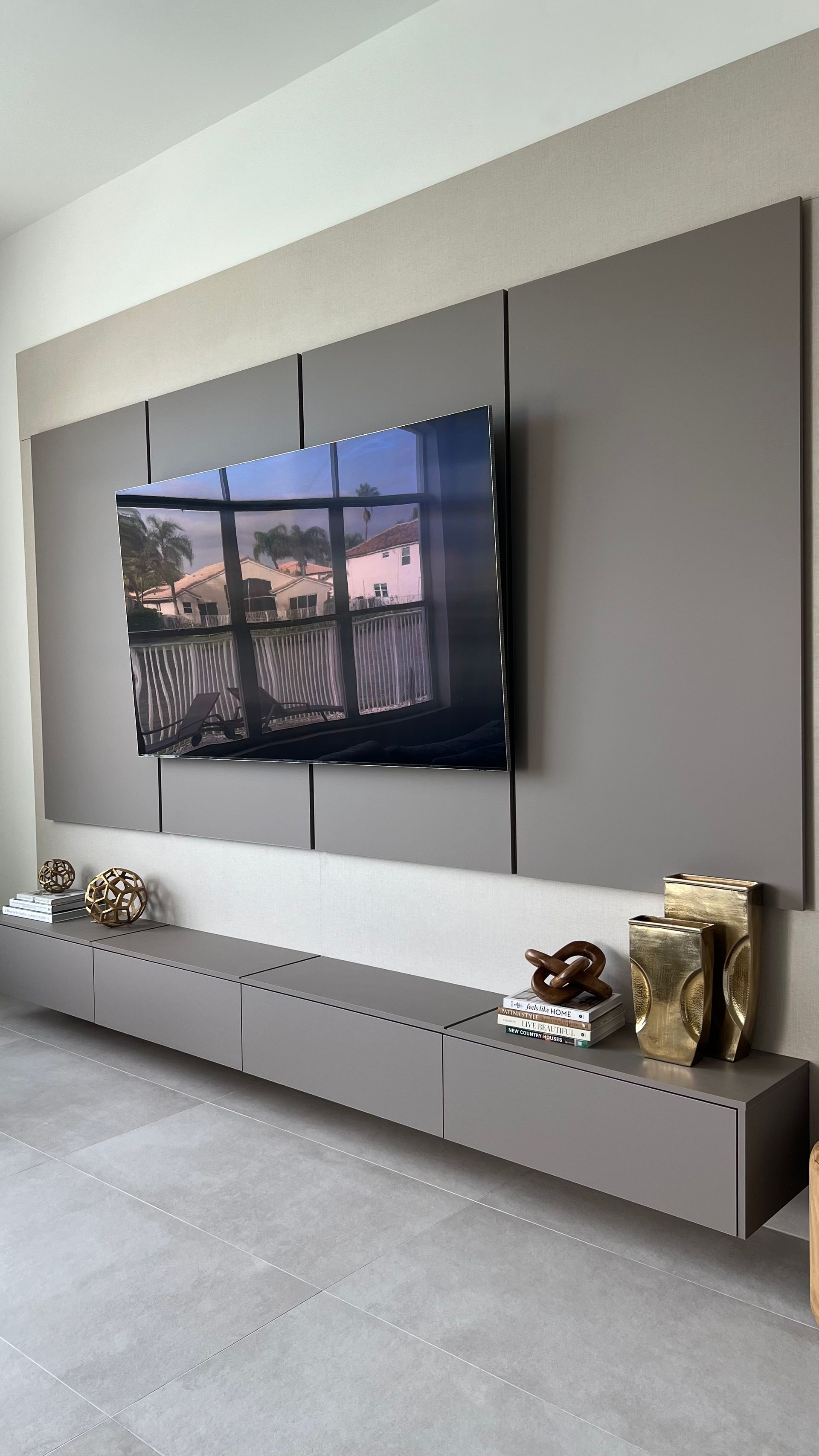 A wall-mounted TV sits above a long, gray floating console with decorative gold accents on a light-colored tile floor.