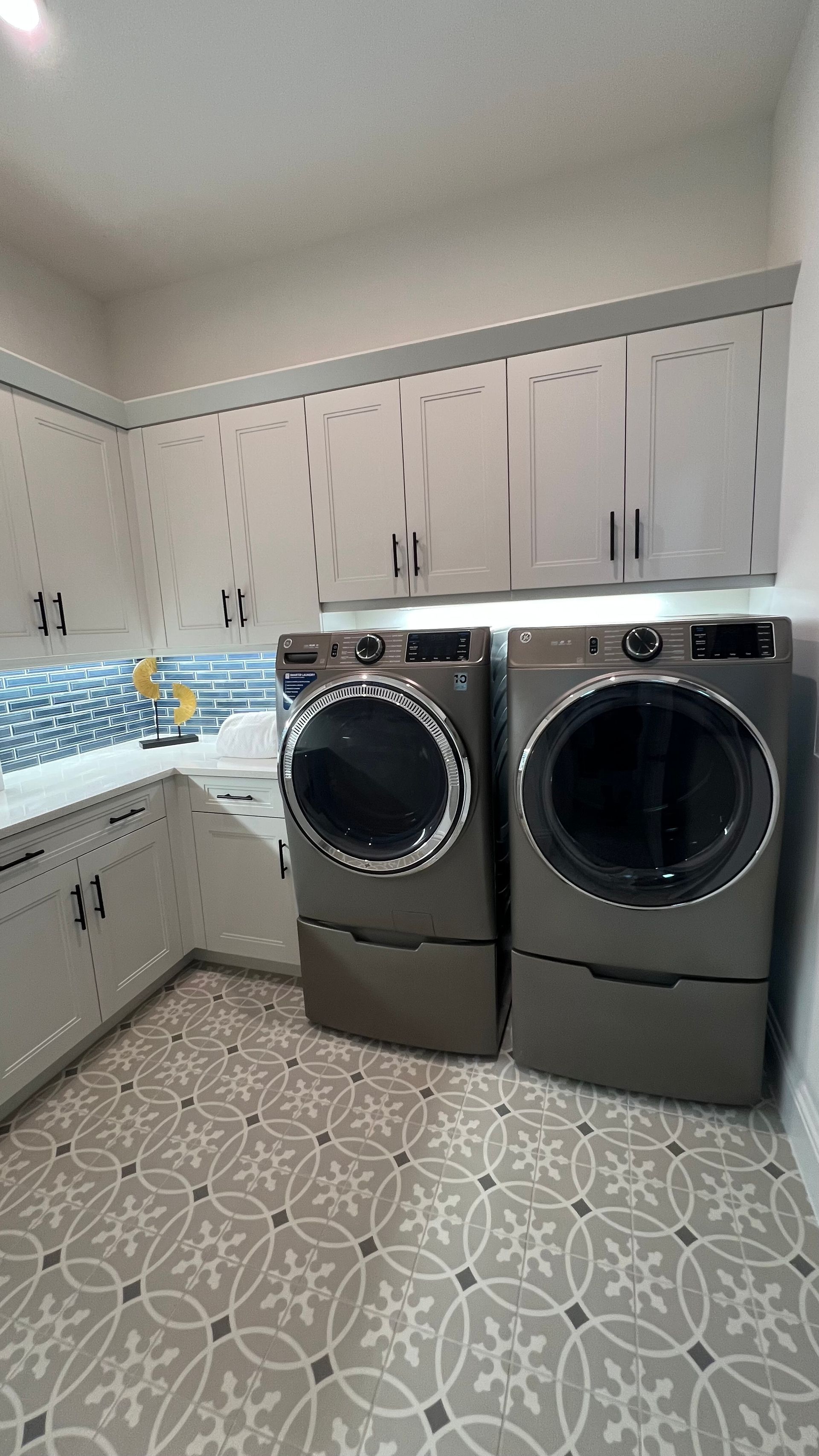 A laundry room with grey cabinets, patterned tile floors, a white countertop, and two dark grey appliances.