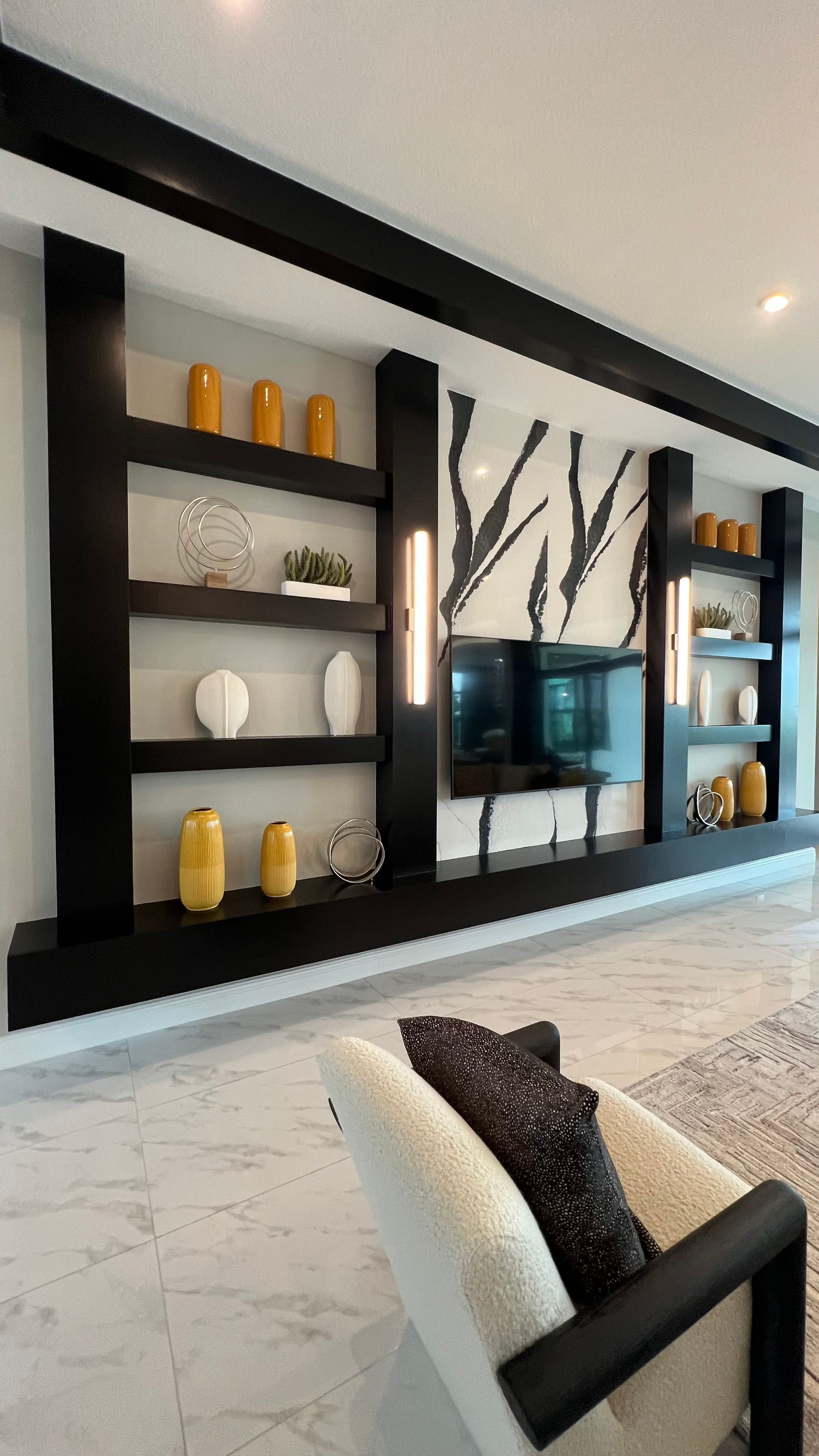 A modern living room wall unit with dark wooden shelving, yellow vases, a flat-screen TV, and a white upholstered chair.