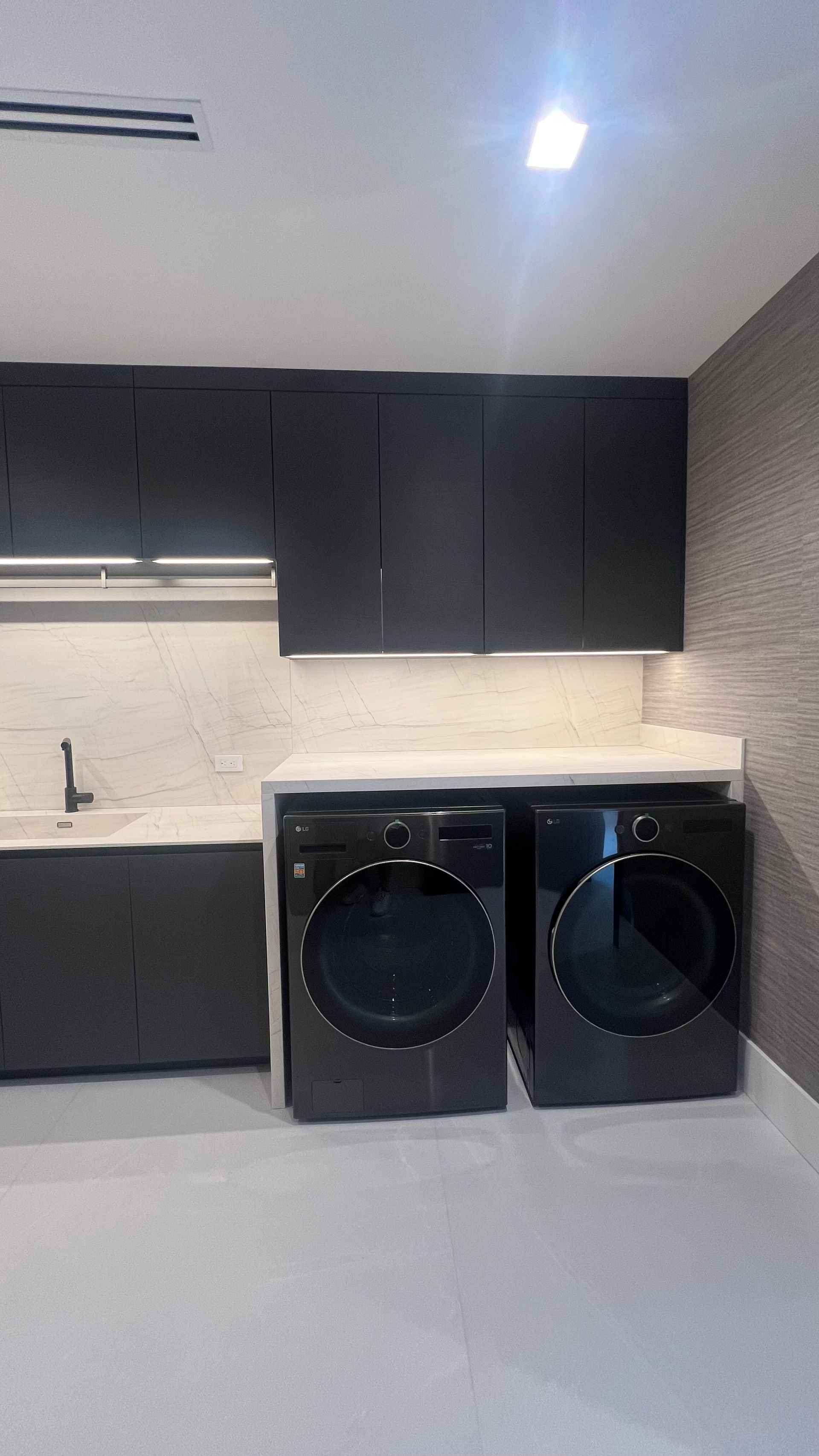 A laundry room featuring modern dark grey cabinetry, a light stone countertop, a sink, and two black washing machines.