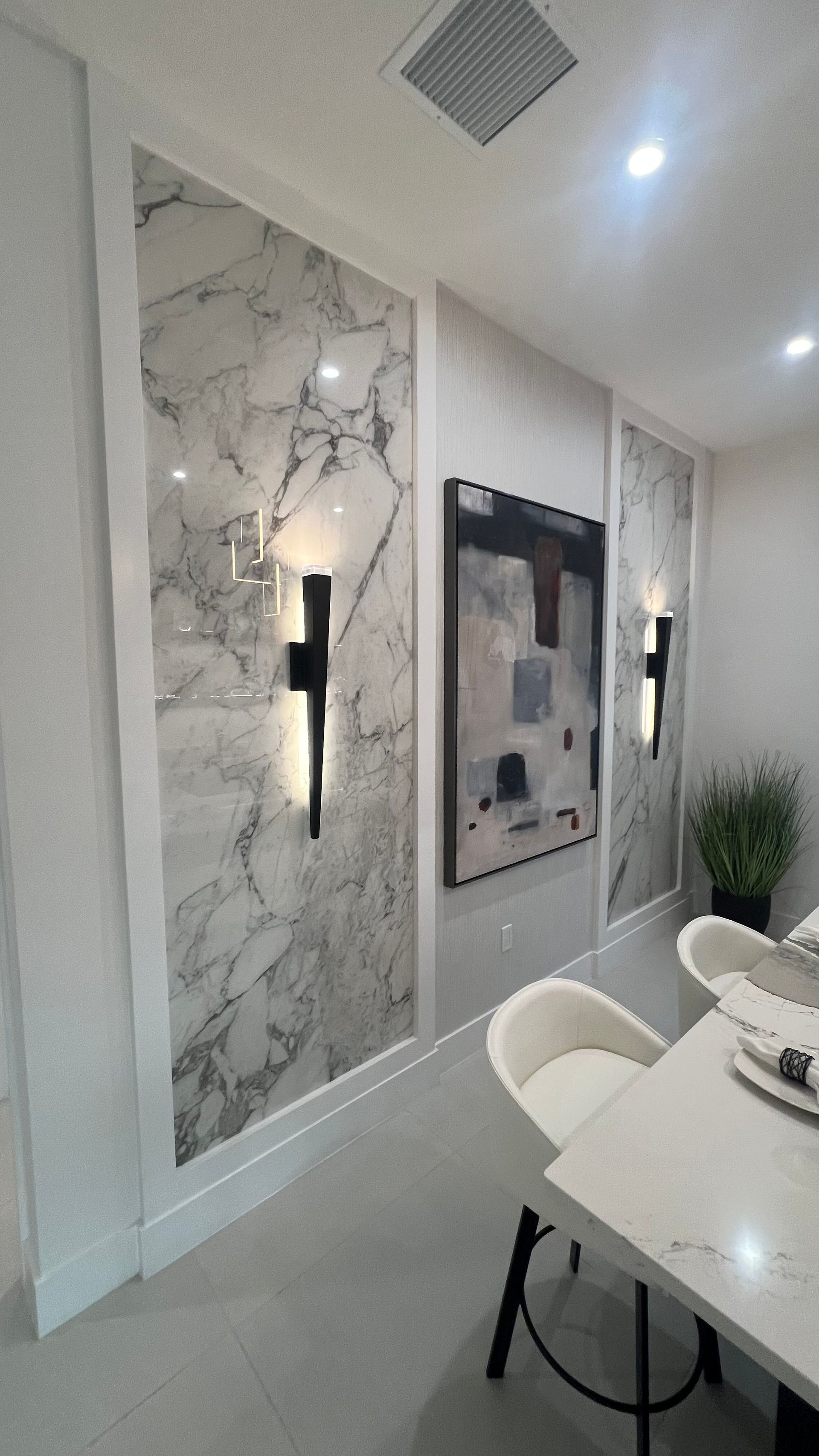 Dining area with two marble wall panels, modern black wall sconces, a framed abstract painting, and white bistro chairs.