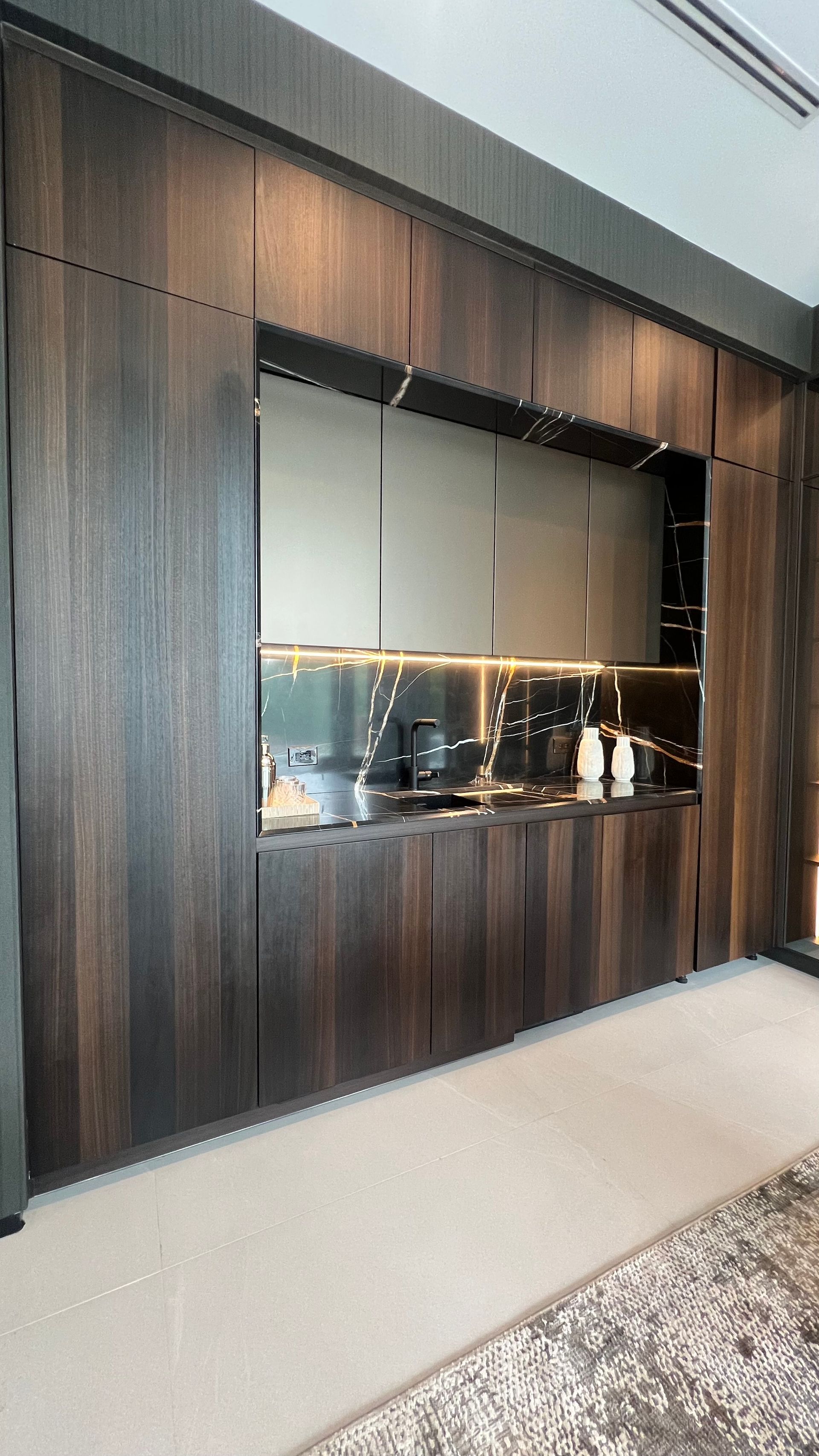 A modern, dark wood kitchen unit featuring a recessed workspace with light gray cabinets, gold trim, and under-cabinet lighting.