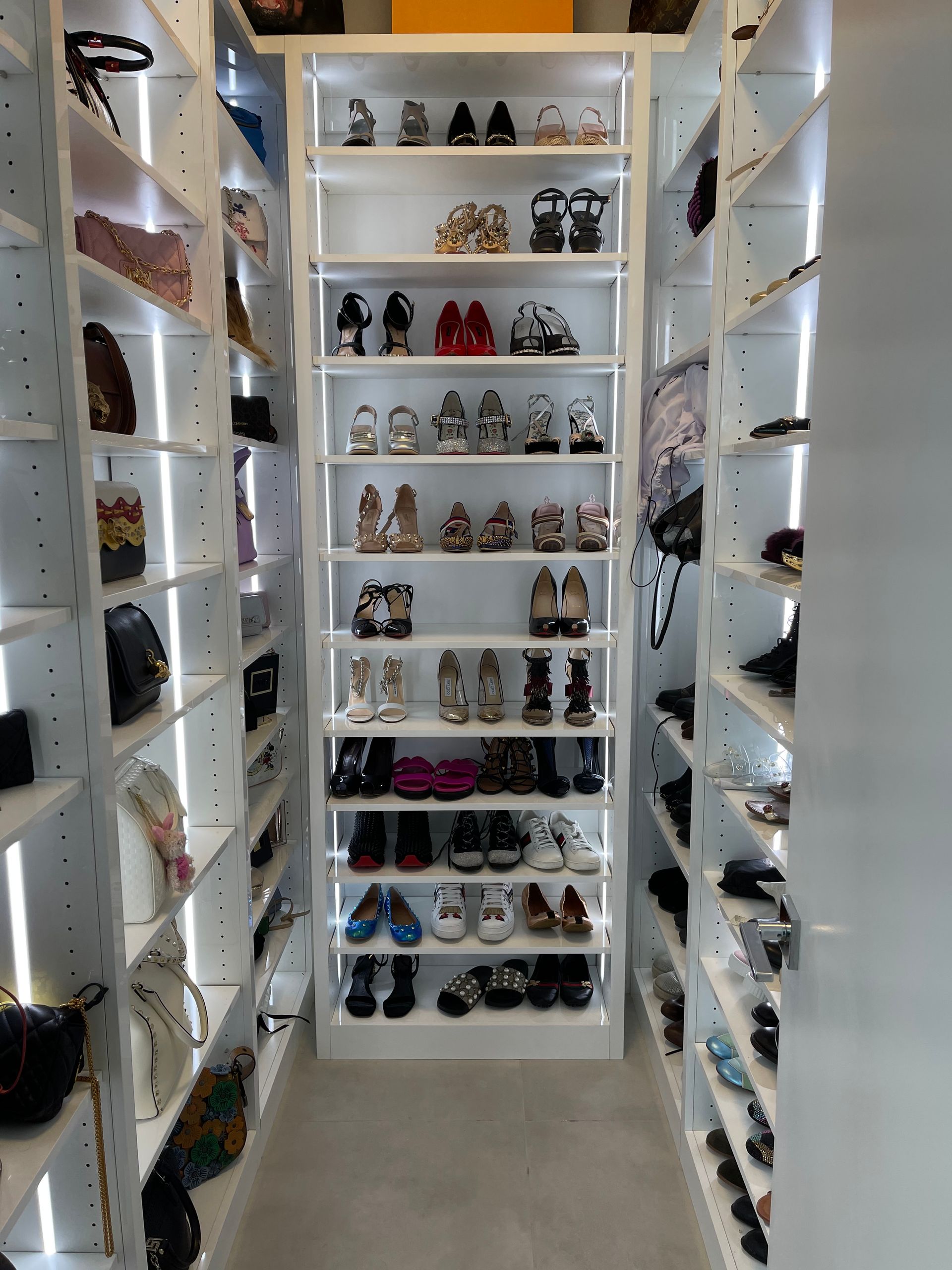 A walk-in closet with illuminated white shelving units filled with various pairs of shoes and handbags.