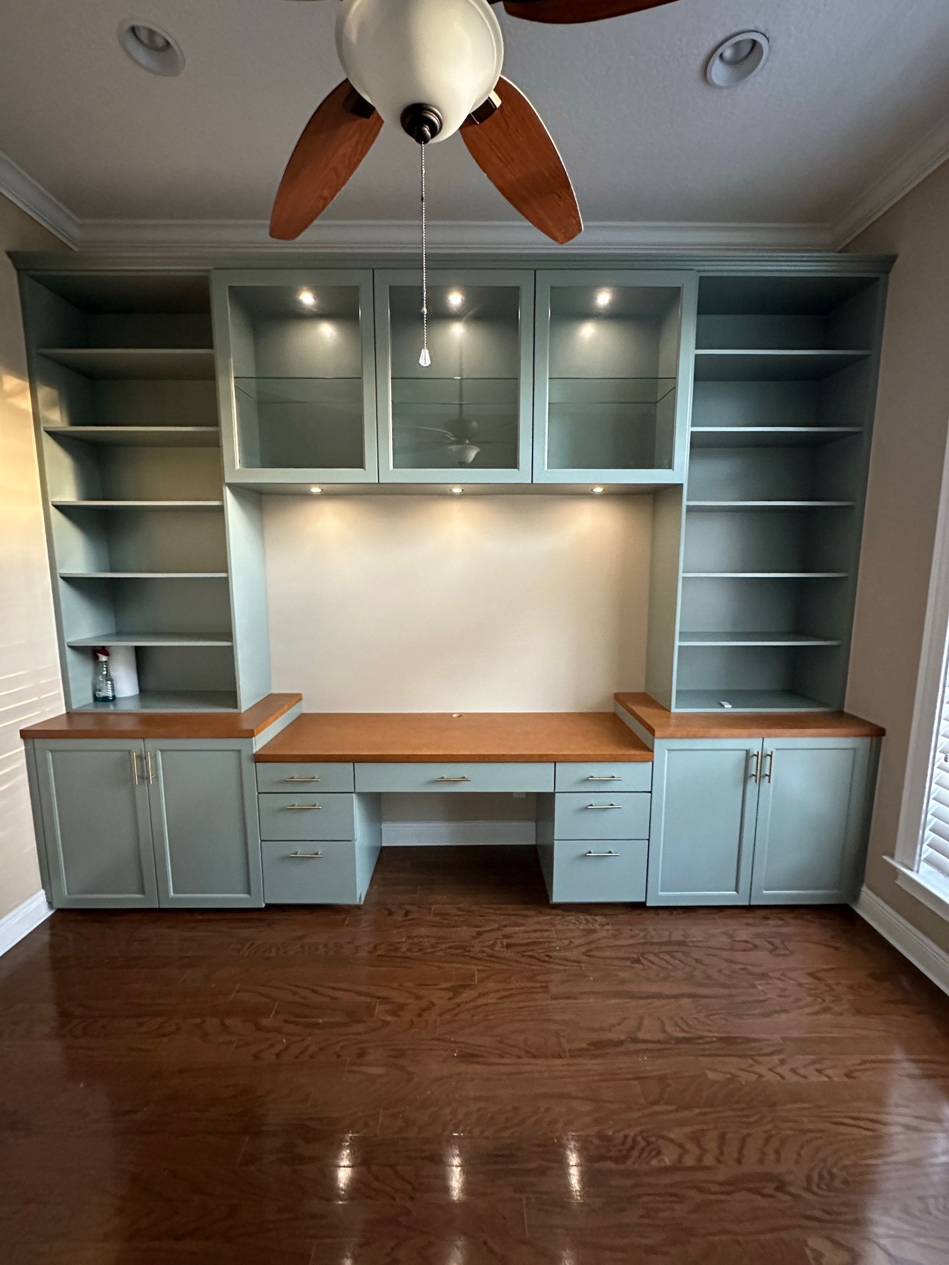 A blue built-in office desk and shelving unit with glass-front cabinets and integrated lighting on a wood floor.