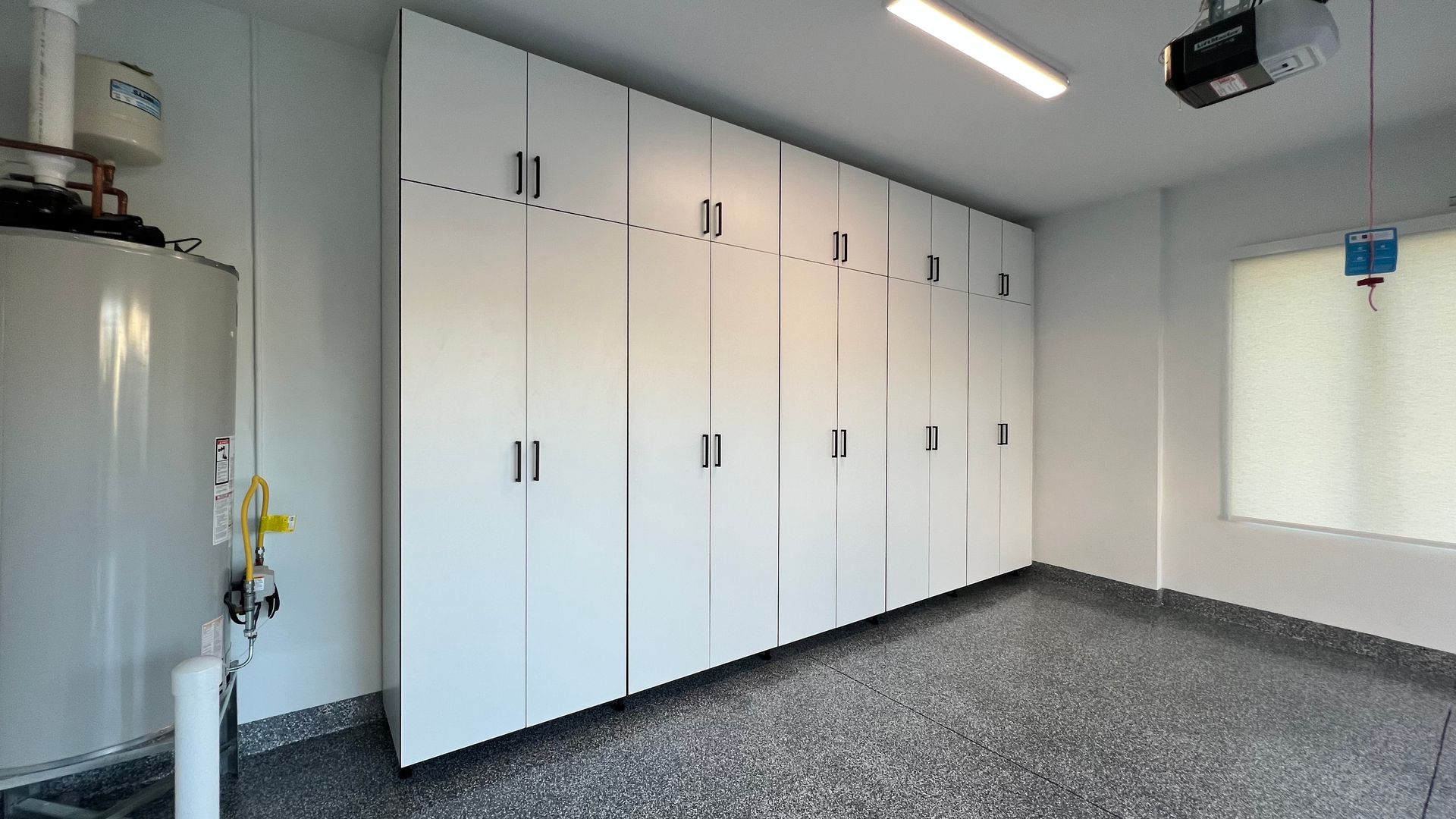A wall of white floor-to-ceiling storage cabinets in a garage with speckled gray flooring and a water heater.