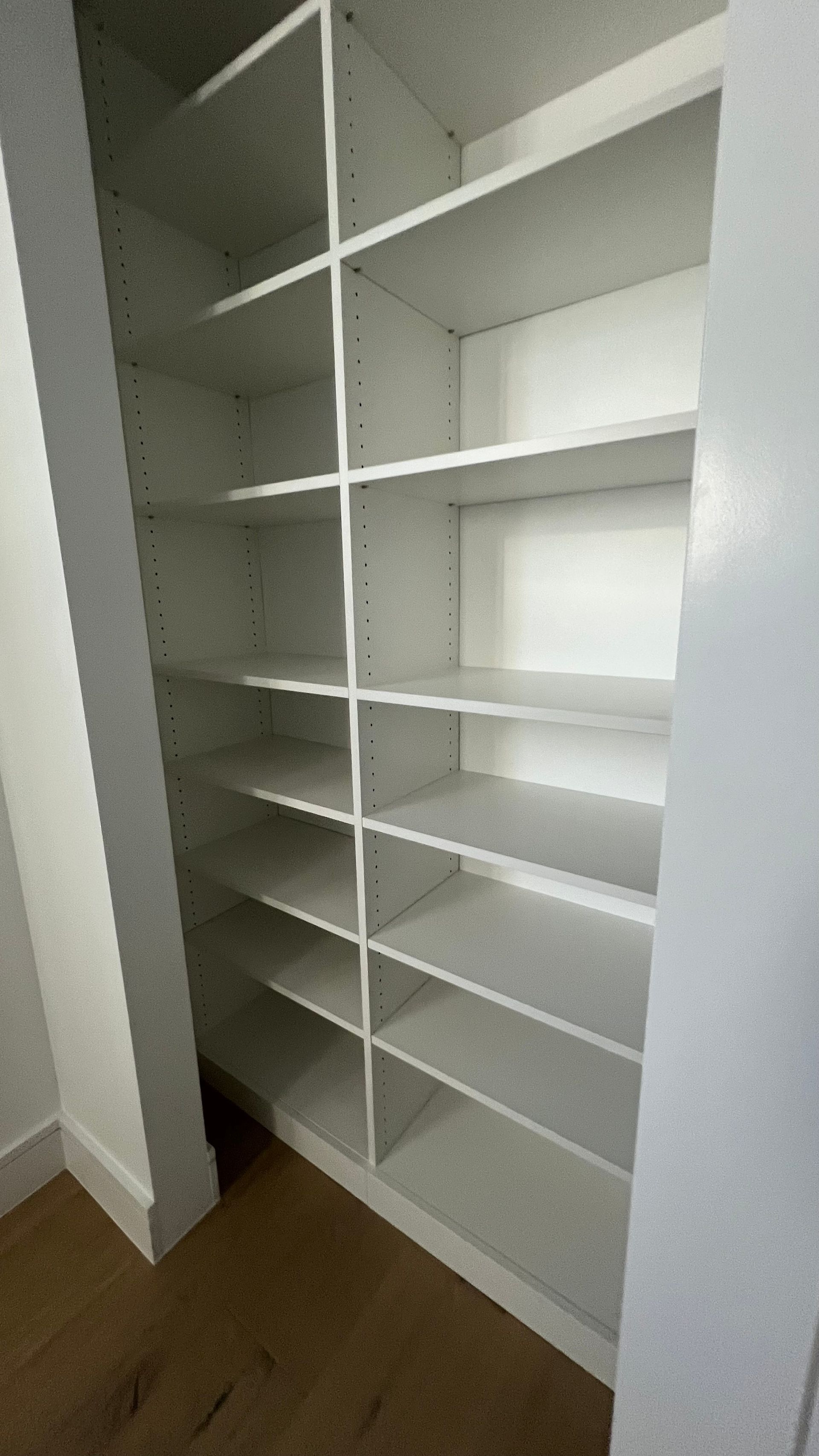 A white, built-in shelving unit with multiple compartments installed in a closet with hardwood flooring.